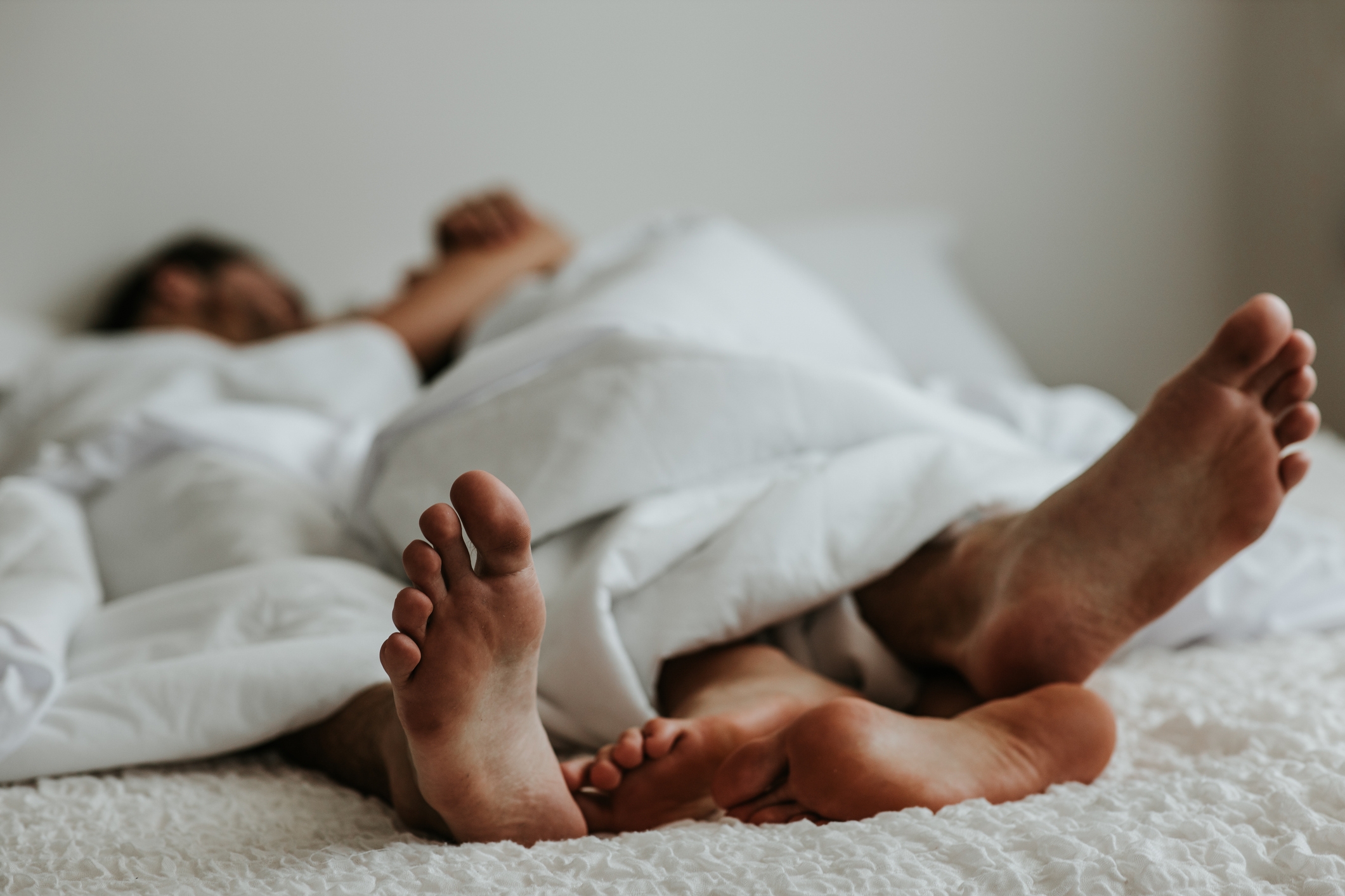 Two people lying in bed with their feet sticking out from under a white blanket. The scene suggests relaxation and comfort