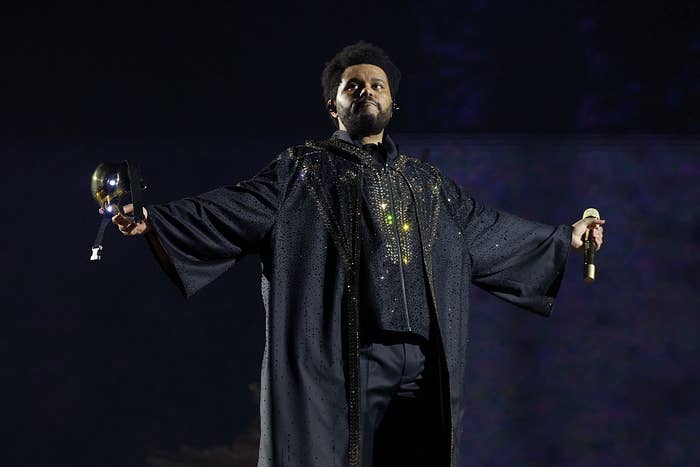 Man on stage in a sparkling robe with wide sleeves, holding a microphone and another object, arms outstretched, performing passionately