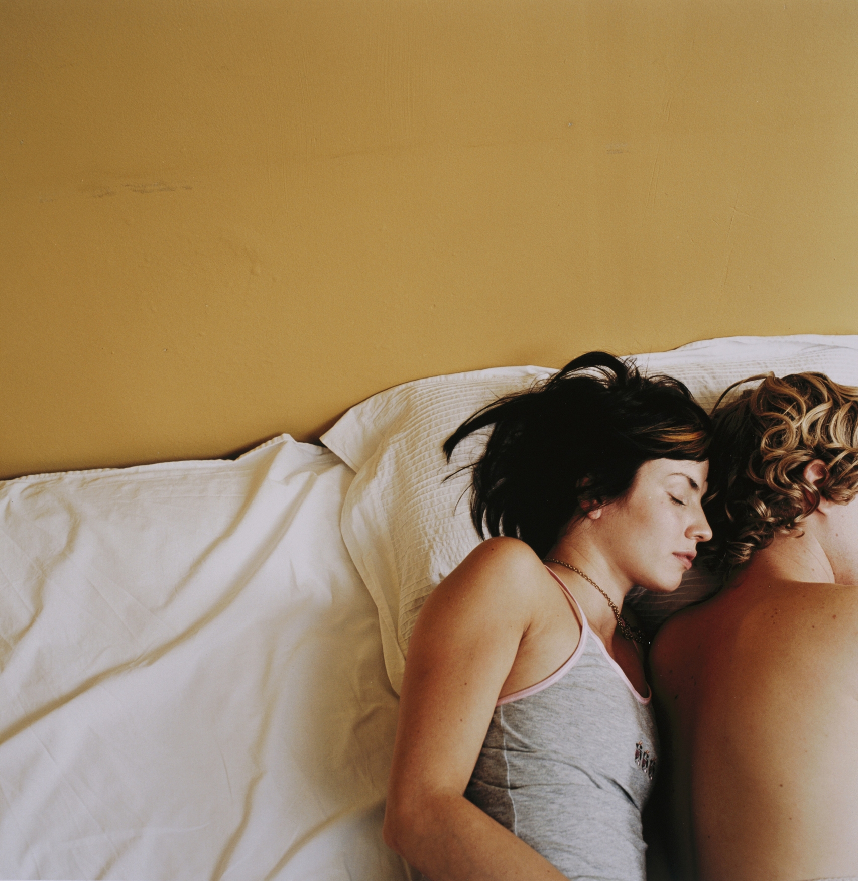 Two people are lying back-to-back on a bed, appearing relaxed. One has long hair and the other's short hair is barely visible