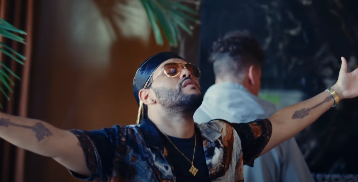 Person with sunglasses and open arms, wearing a patterned shirt and pendant necklace. Another person is in the background