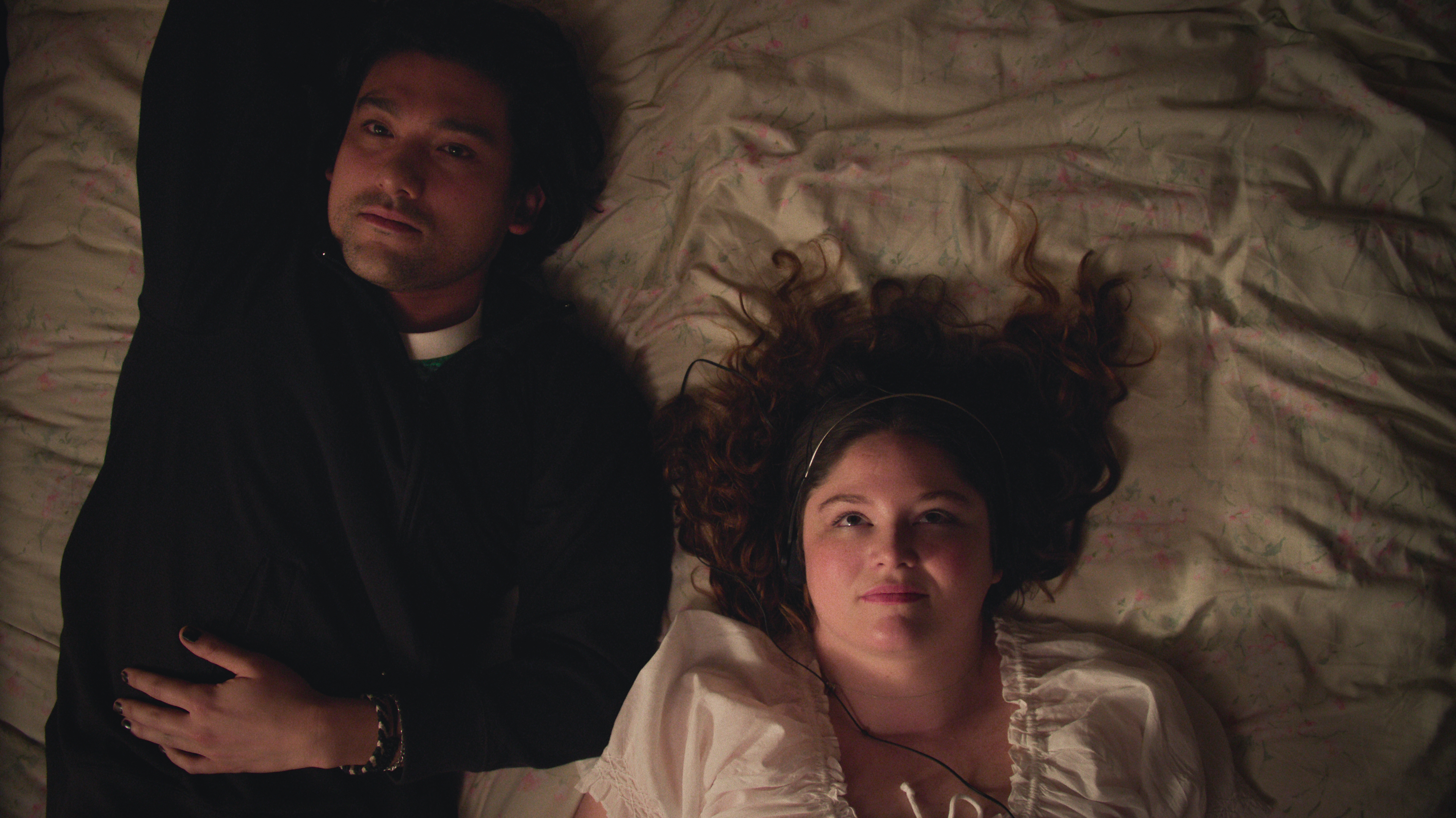 Two people lying on a bed, one wearing a dark top and the other in a light blouse, looking thoughtful