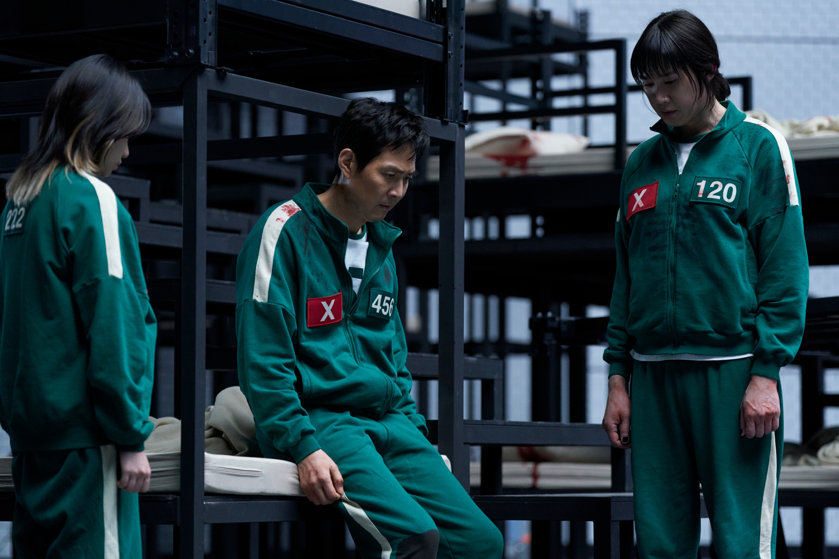 Actors in numbered tracksuits stand in a tense scene from a dramatic TV series, set in a room with metal bunk beds