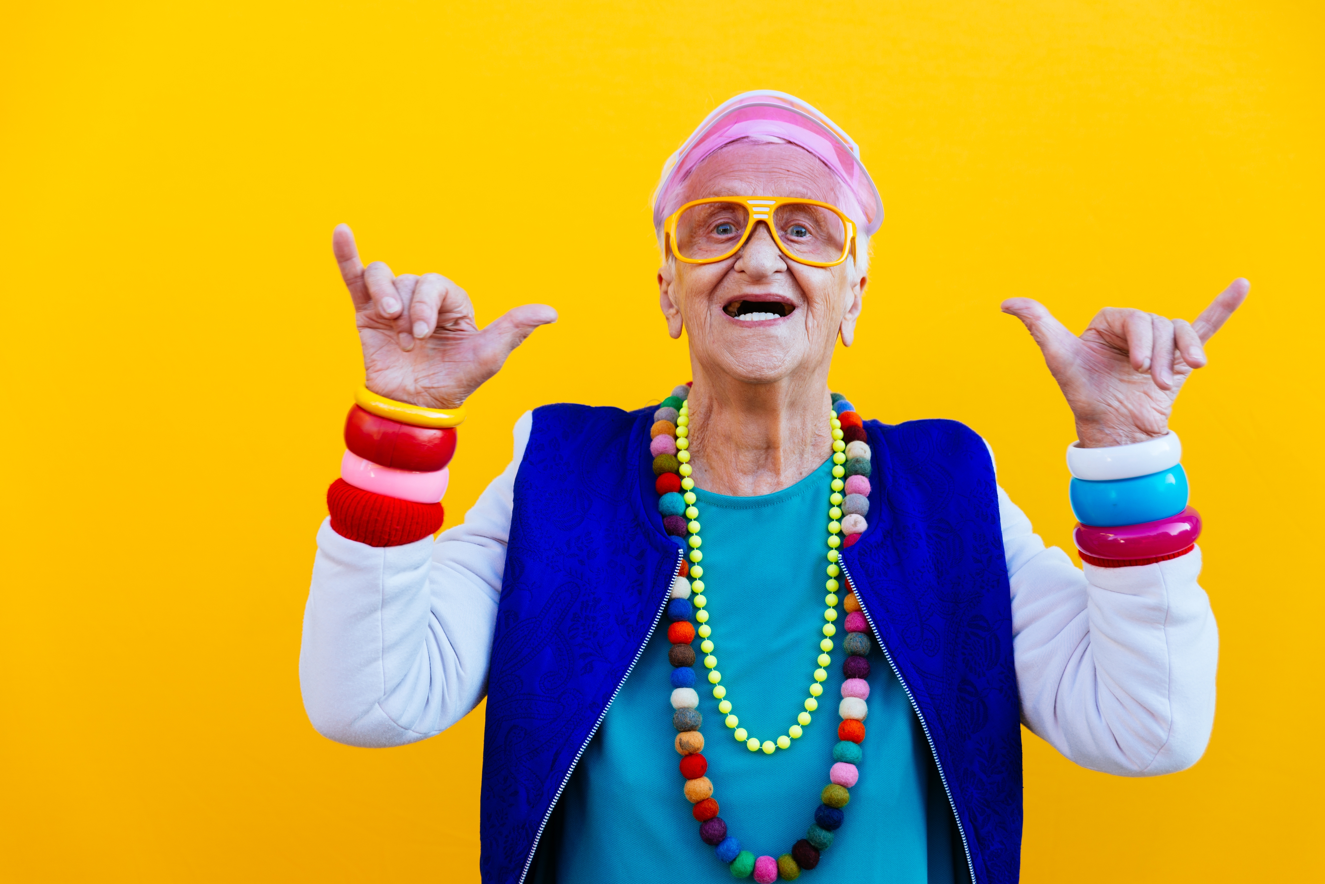 Elderly person joyfully posing with raised pinky fingers, wearing vibrant accessories, including necklaces and bracelets, against a plain background