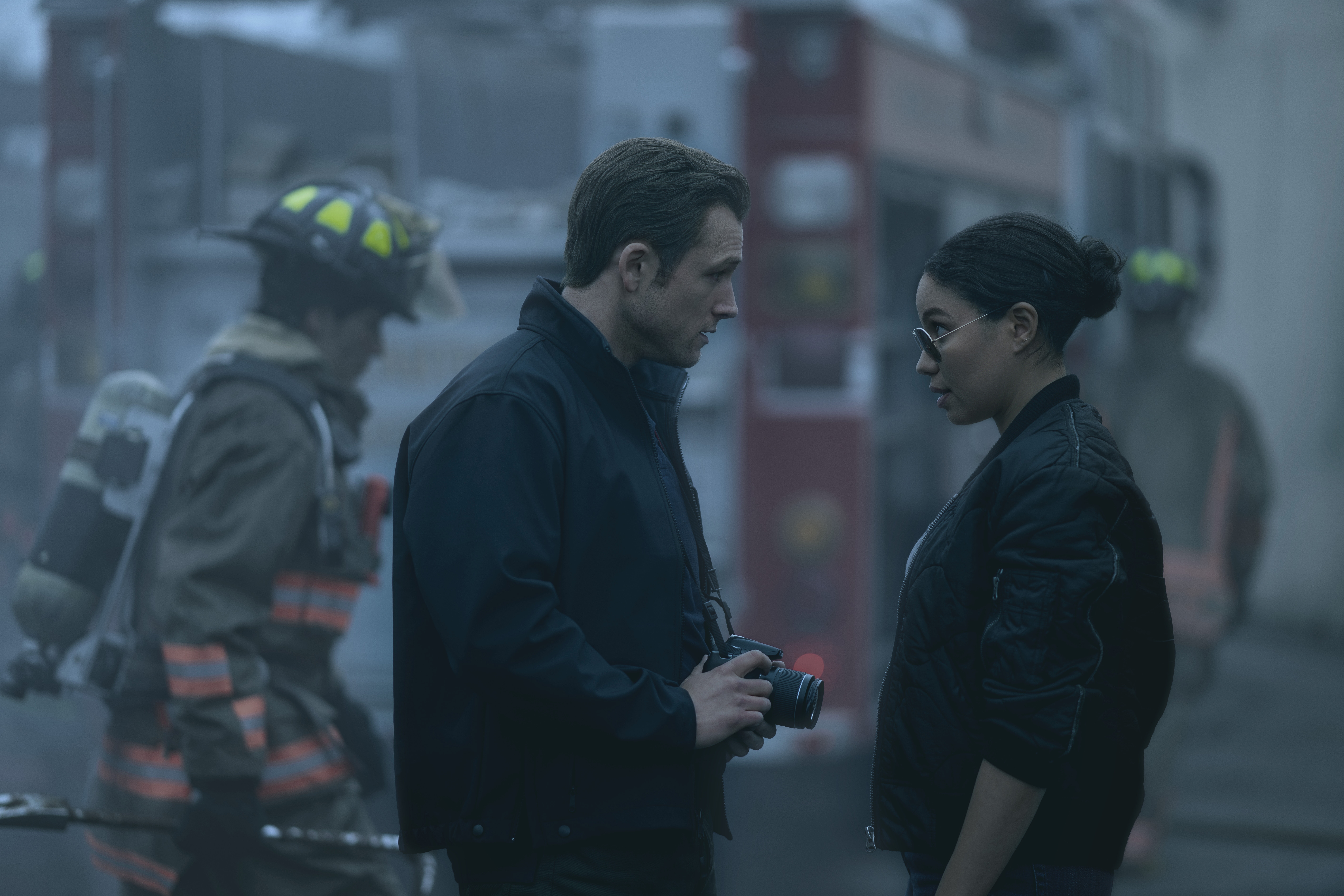 Two people with serious expressions stand near firefighters and a fire truck. One holds a camera, hinting at a tense or investigative scene