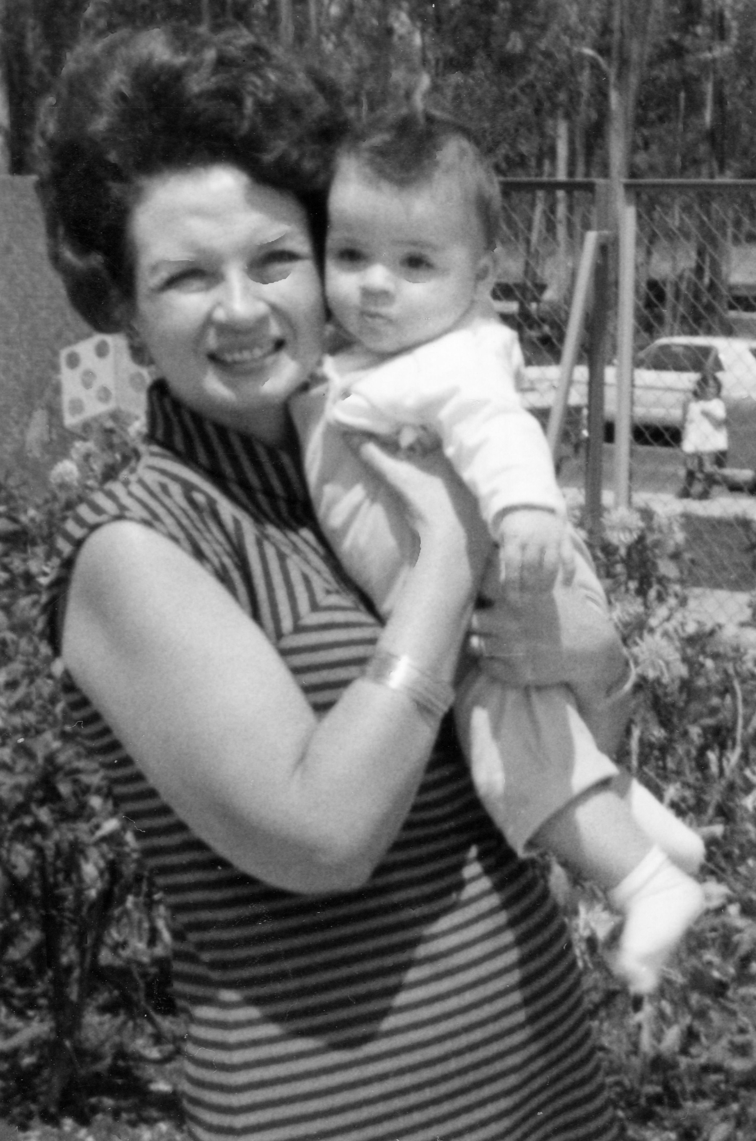 Woman in a striped dress holds a baby outdoors, both smiling. The background features a car and trees
