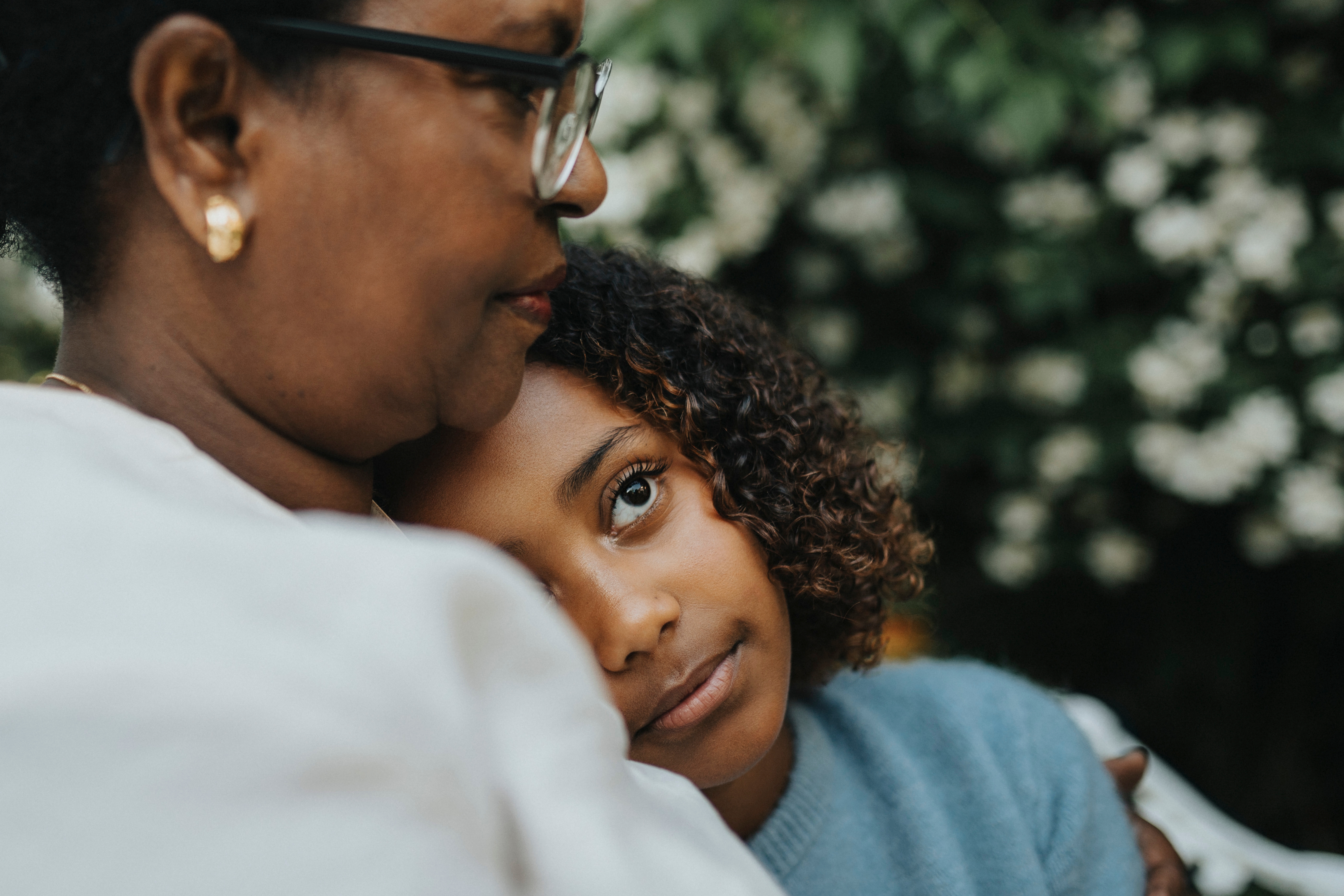 A child rests their head on an older adult's shoulder, looking thoughtfully upward. They are outdoors with greenery in the background