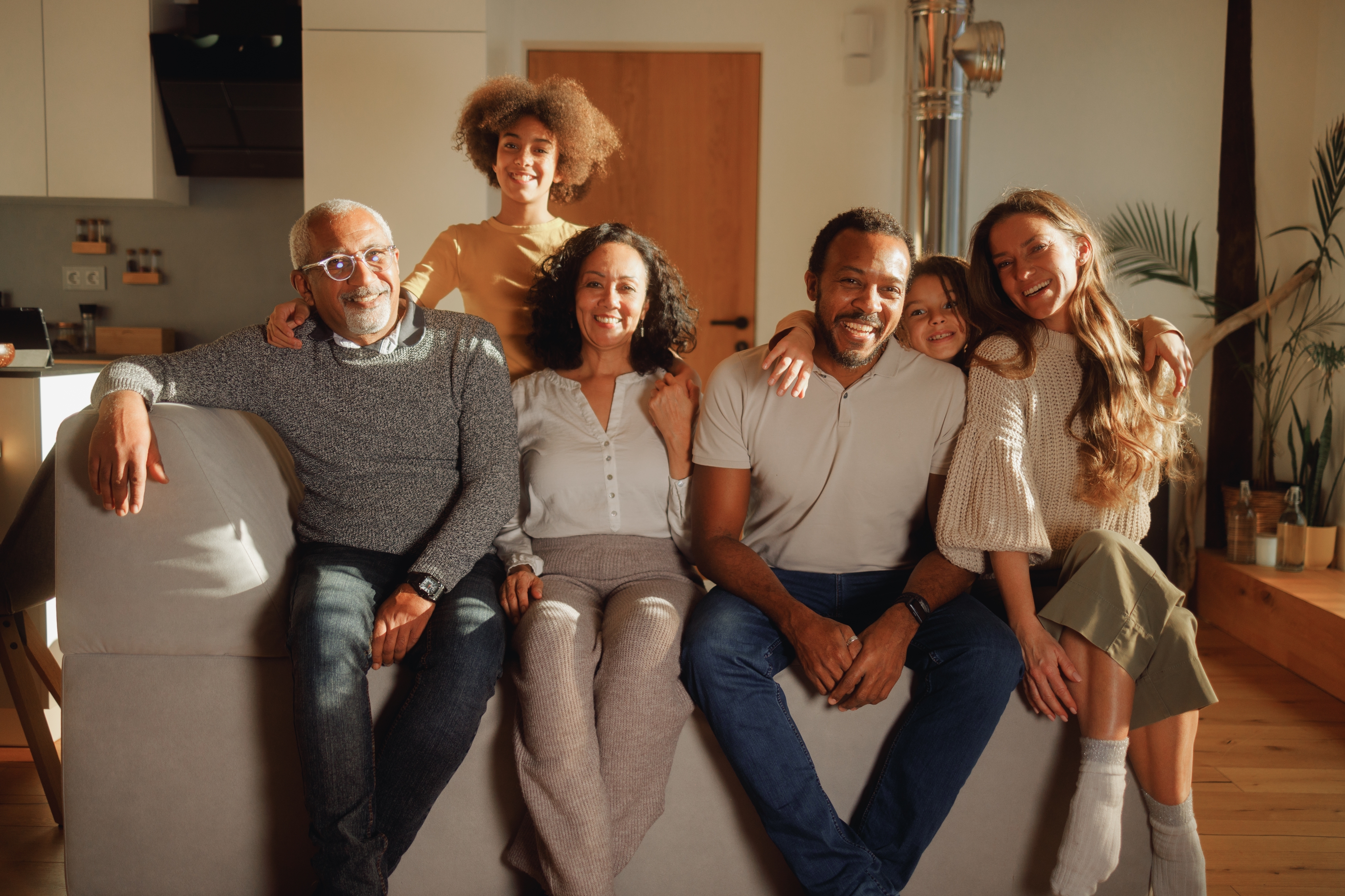 A smiling family of six, including an elderly couple, poses together casually on a sofa in a bright living room