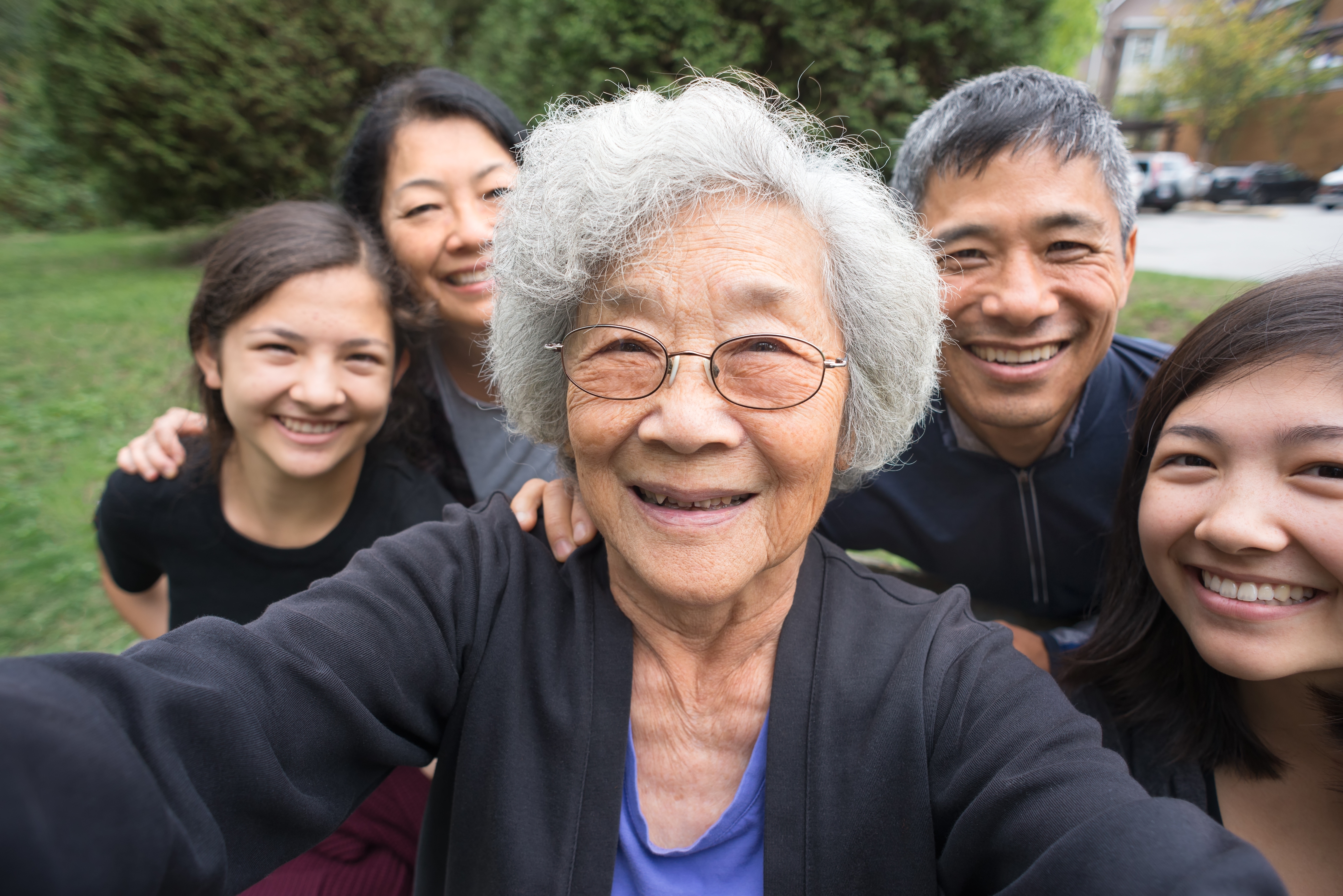 A joyful elderly woman takes a selfie with her smiling family outside in a park, capturing a moment of togetherness