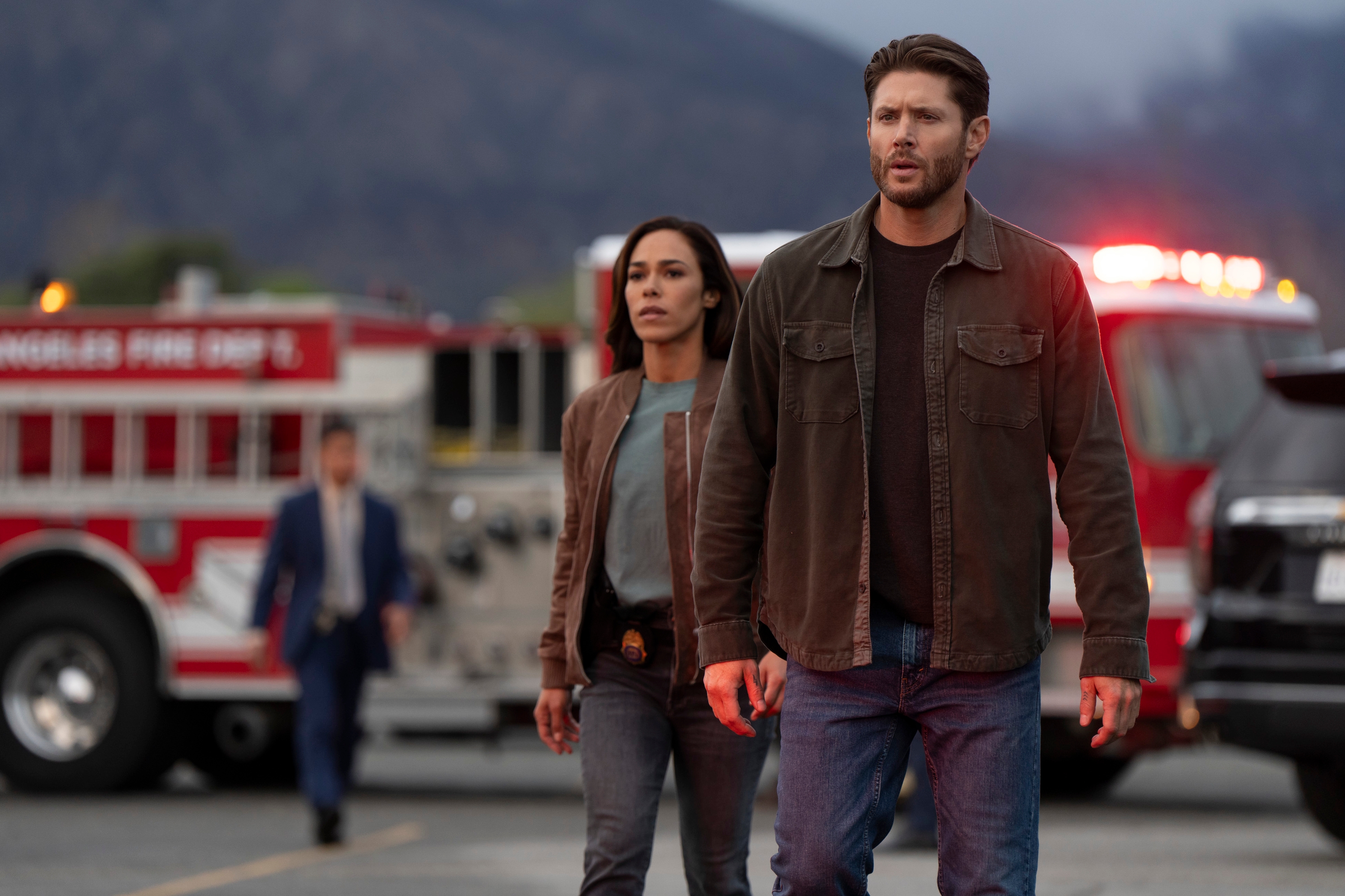 Two actors in casual jackets and jeans walk with a serious expression in front of blurry fire trucks and emergency lights in the background