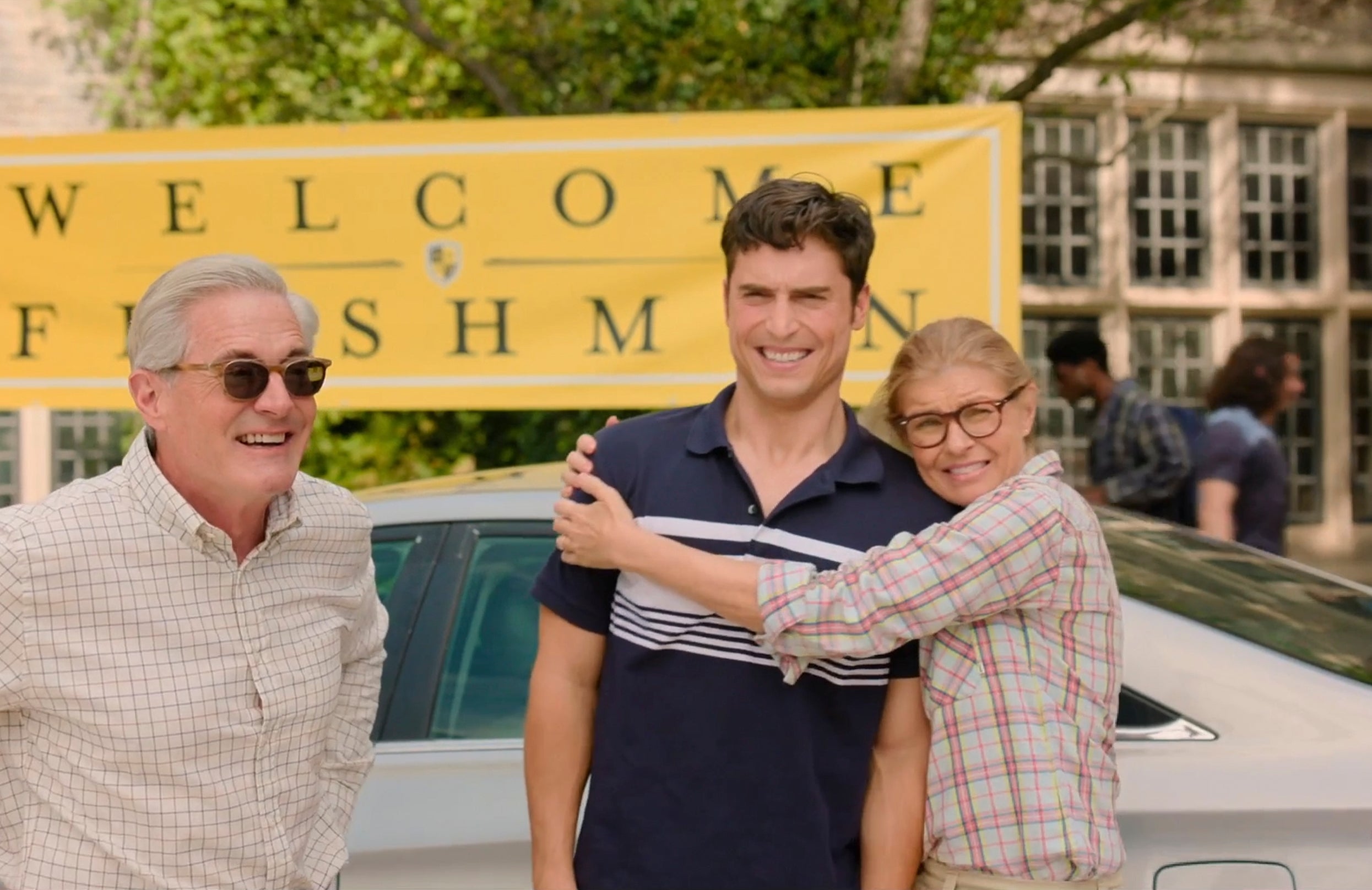 A smiling young man stands with two older people embracing him in front of a "Welcome Freshman" banner at a school campus setting