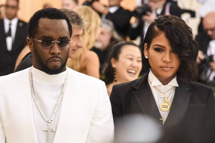 Two celebrities on a red carpet, one in a white suit with necklaces, the other in a tuxedo with layered necklaces, both with serious expressions