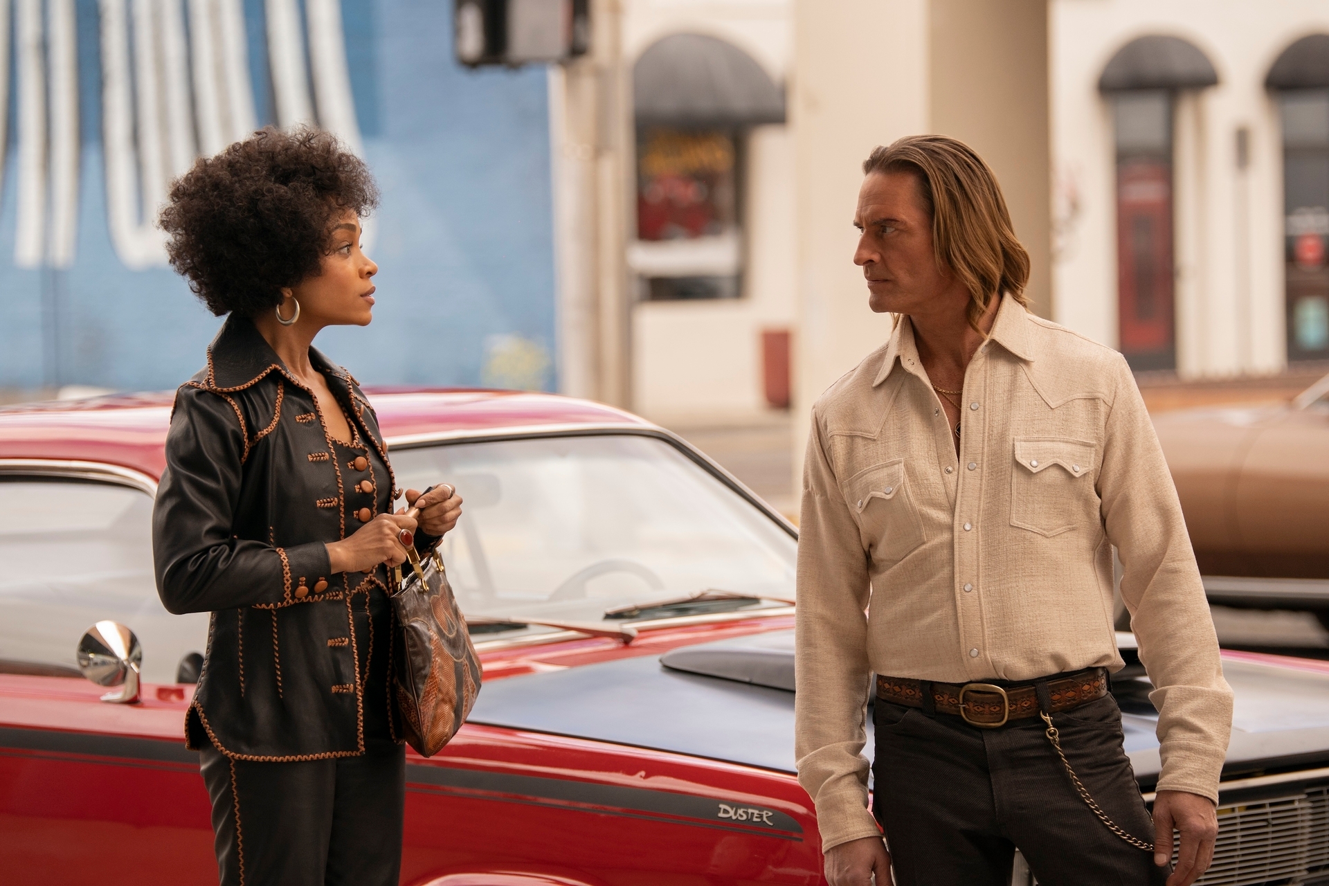 Two actors in a vintage street scene; one in a detailed leather outfit, the other in a button-up shirt and jeans
