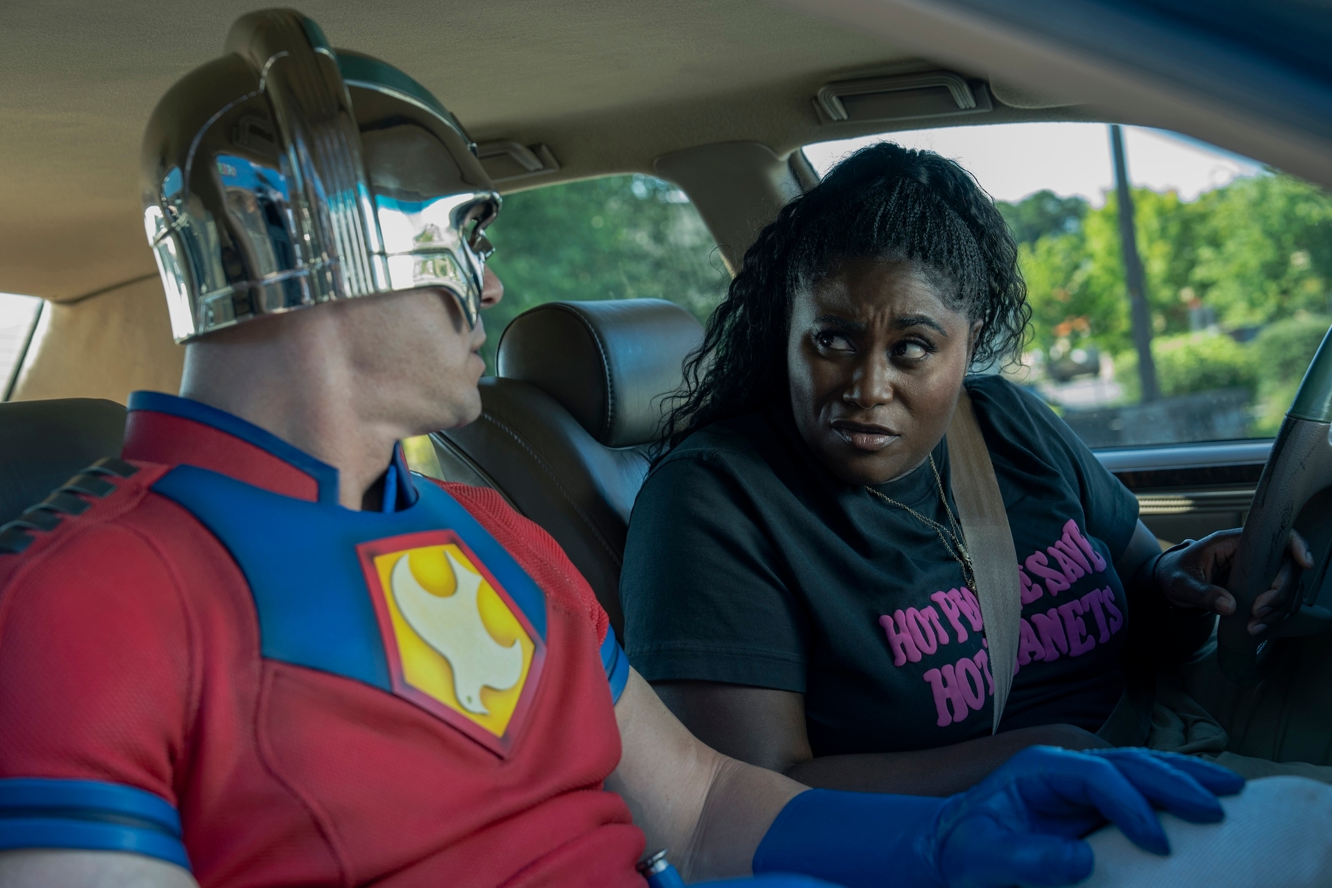 Two characters from a TV show sit in a car. One wears a superhero costume with a helmet; the other wears a casual T-shirt, looking at each other