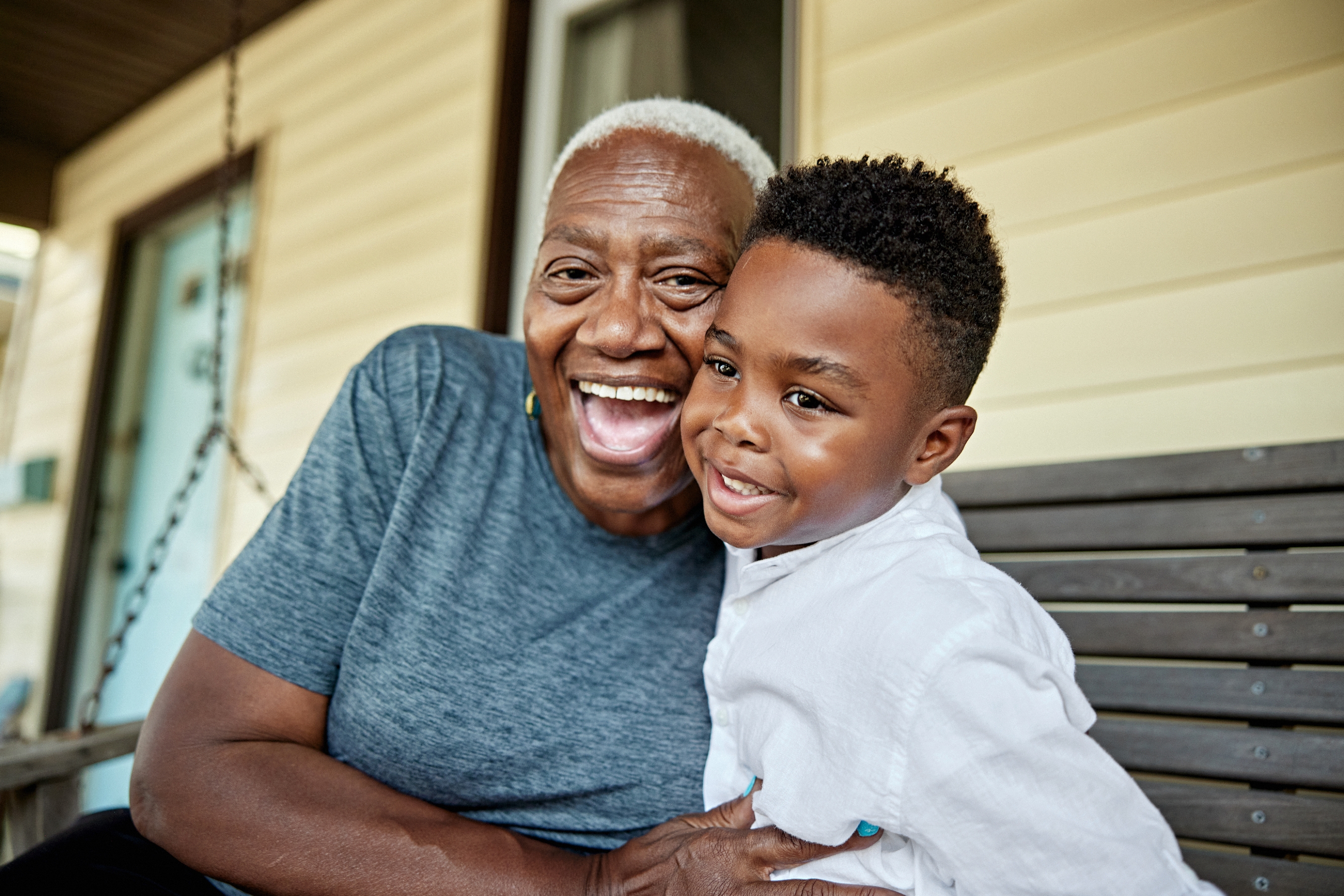 A joyful older adult and a child, sitting on a porch swing, smile warmly at the camera