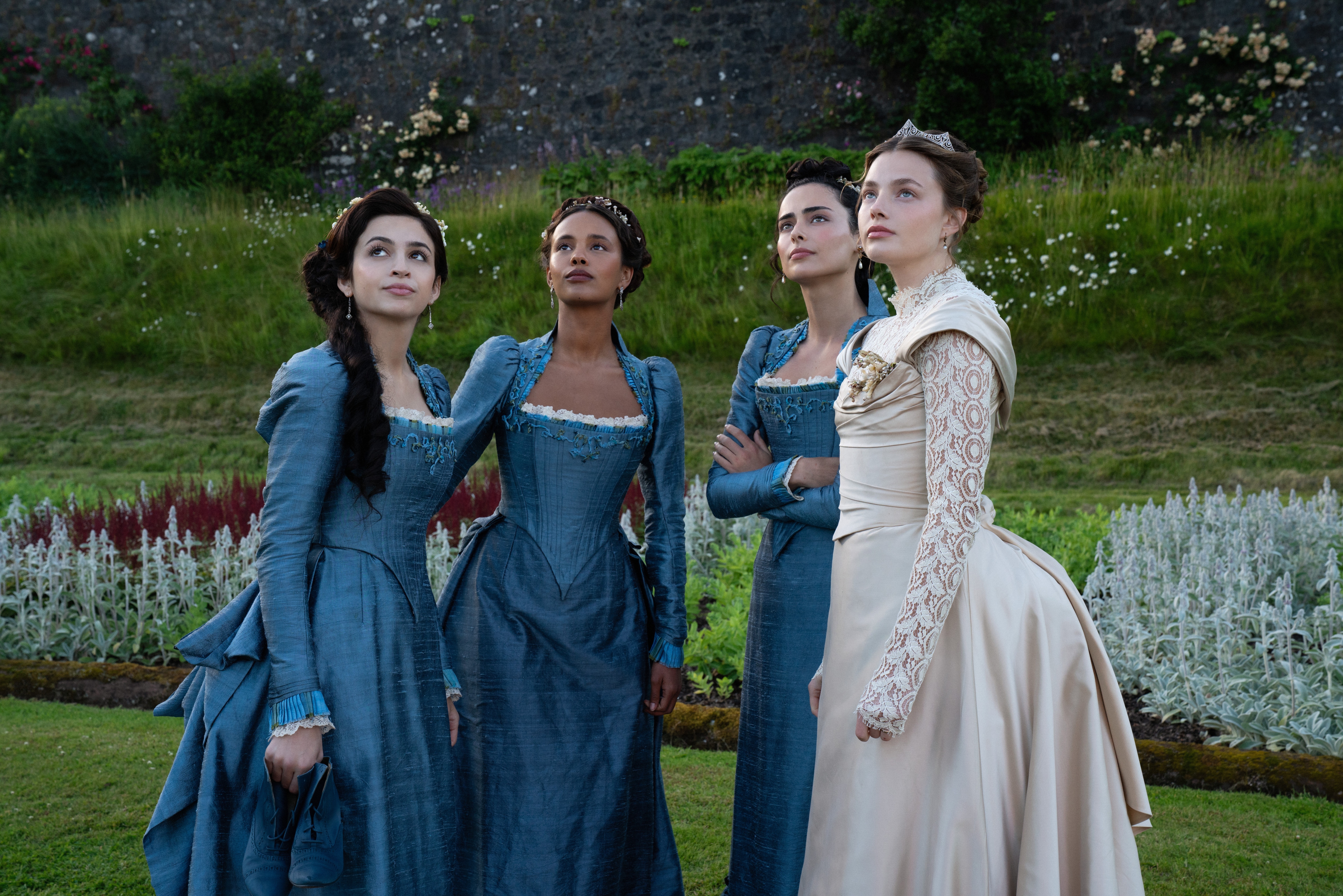 Four women in period costumes, three in blue dresses, one in white, stand outside gazing upwards, appearing as if in a historical TV or movie scene