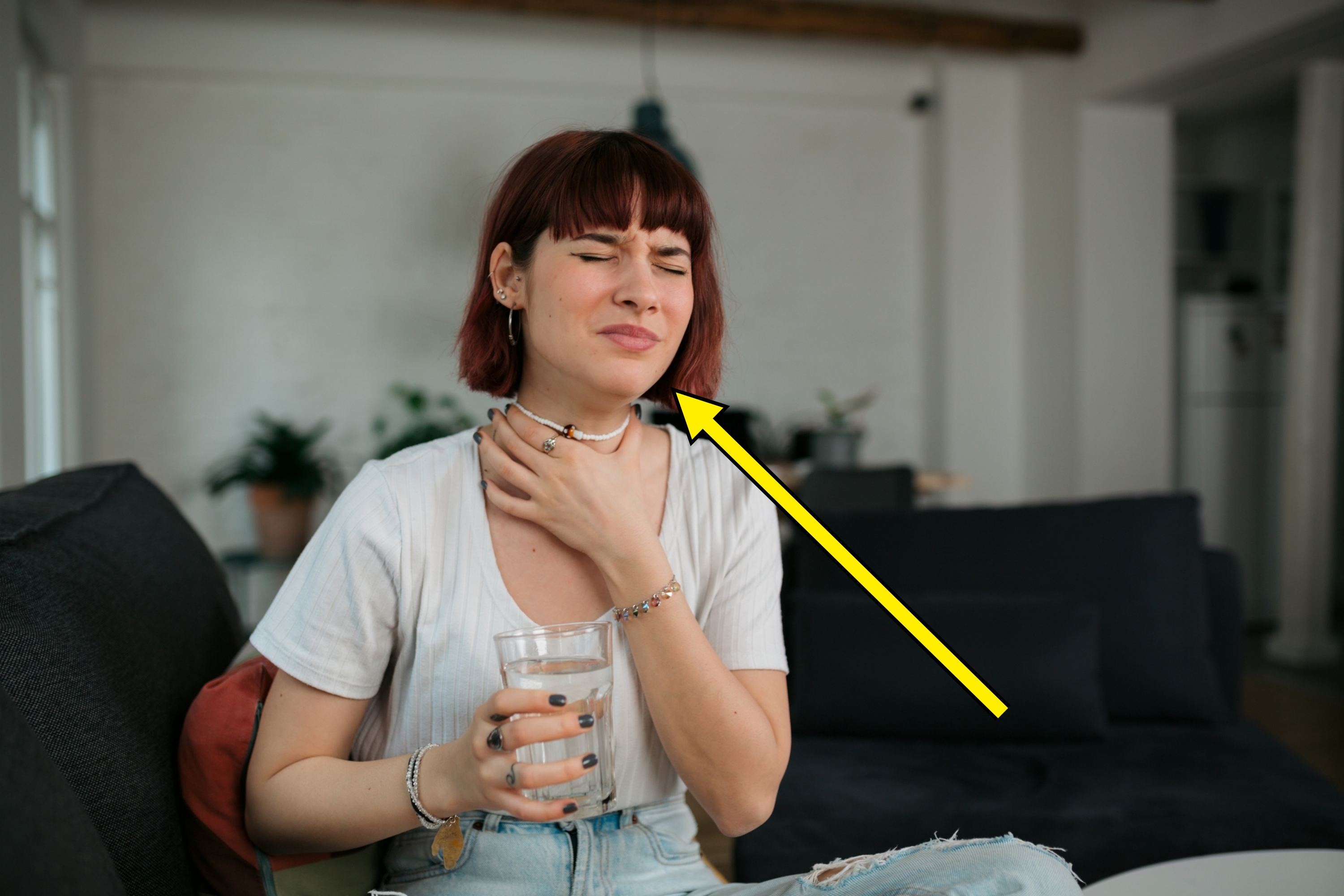 A person sits on a couch holding their throat with one hand and a glass of water in the other, appearing uncomfortable