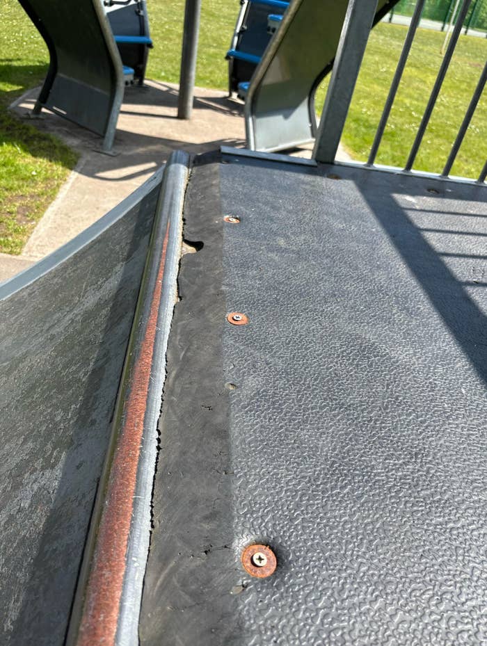 Close-up of a playground slide showing rusted edges and worn surface, with part of another slide and grassy area in the background