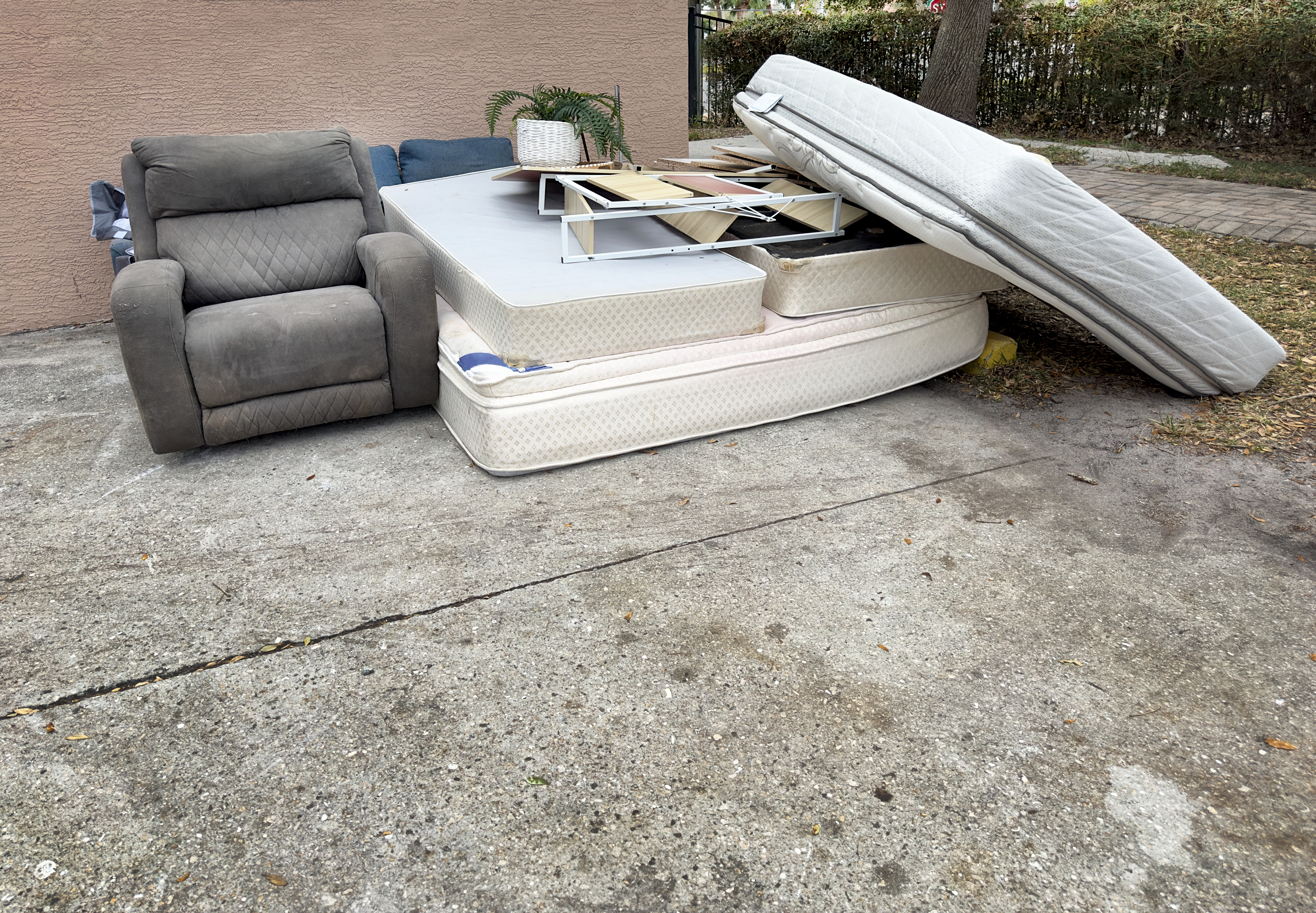 Discarded furniture on street with a gray recliner, mattresses, a table, and a cushion against a wall