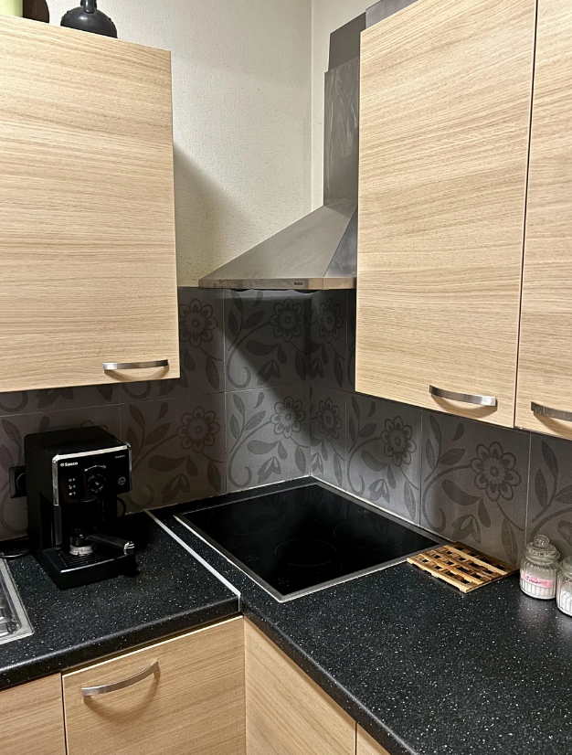 Modern kitchen with wooden cabinets, black countertop, electric stove, range hood, and coffee machine. Floral backsplash design visible
