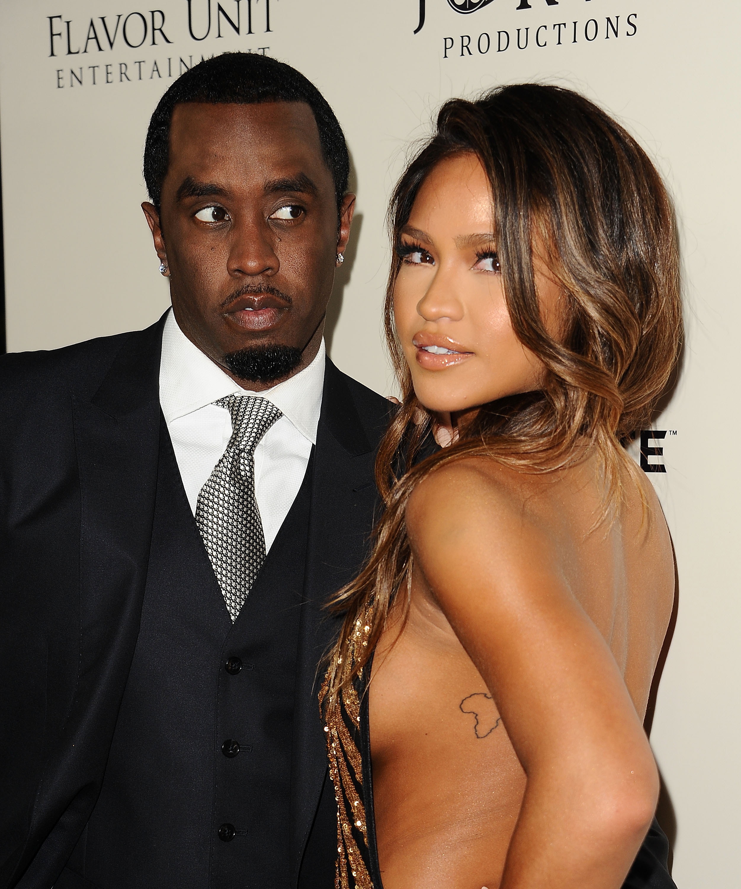 Two celebrities pose on the red carpet. The man wears a suit with a tie; the woman is in a revealing, stylish gown