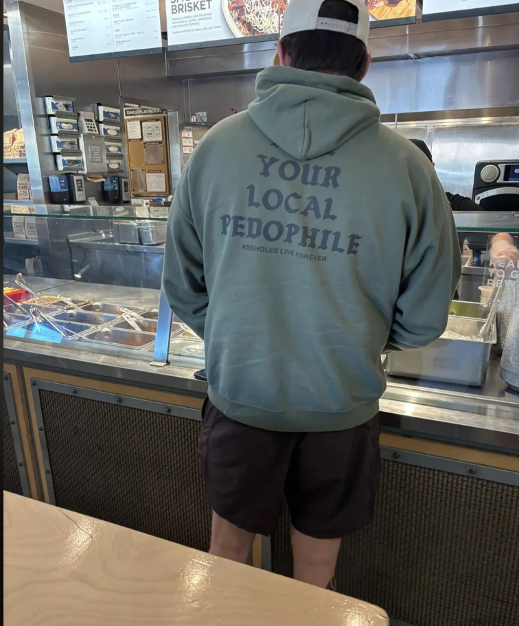A person in shorts and a hoodie with text on the back, standing at a food counter. The text reads, "Your local pedophile."