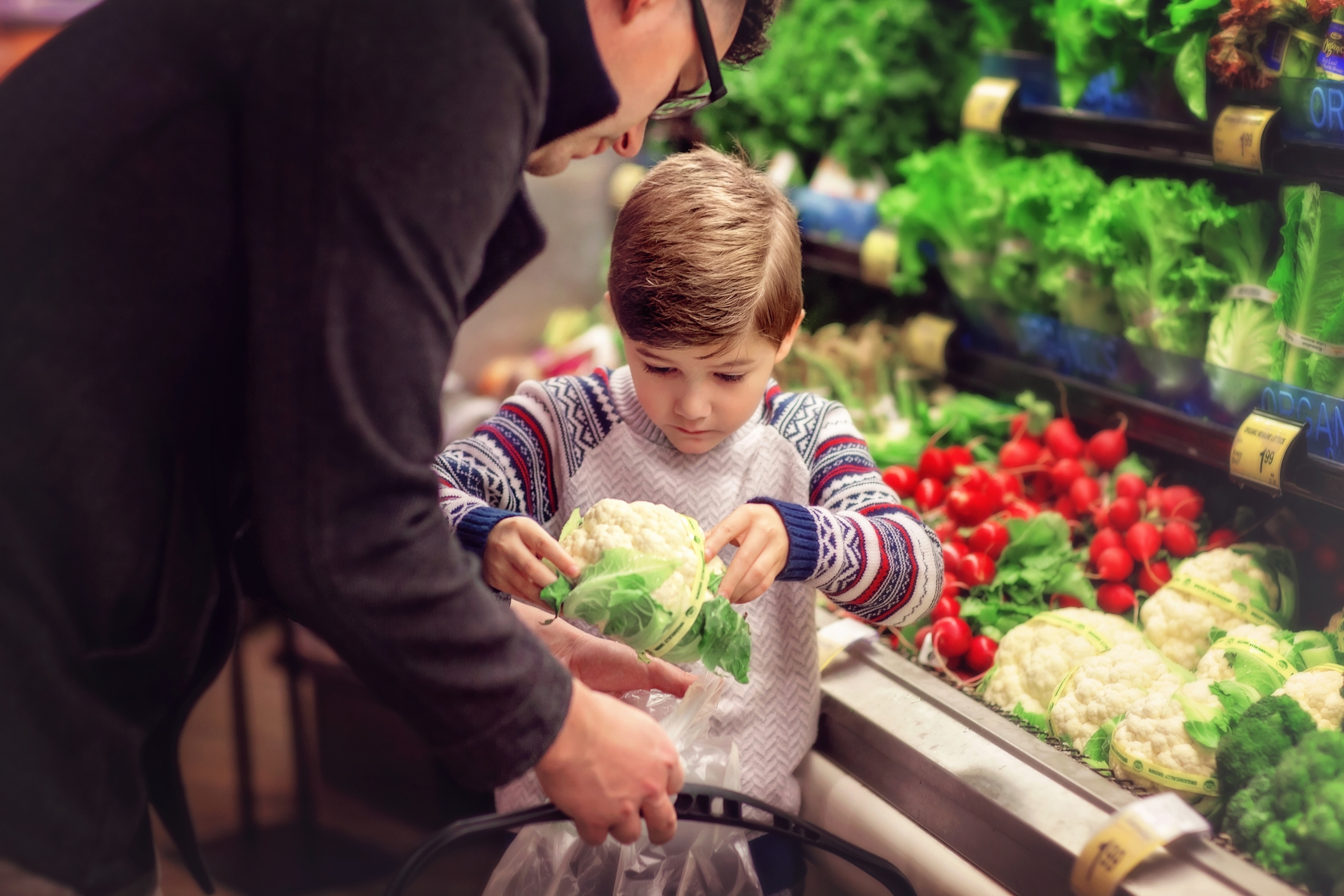 A man helps a child at a grocery store select fresh cauliflower and produce, suggesting a learning moment about produce selection and budgeting