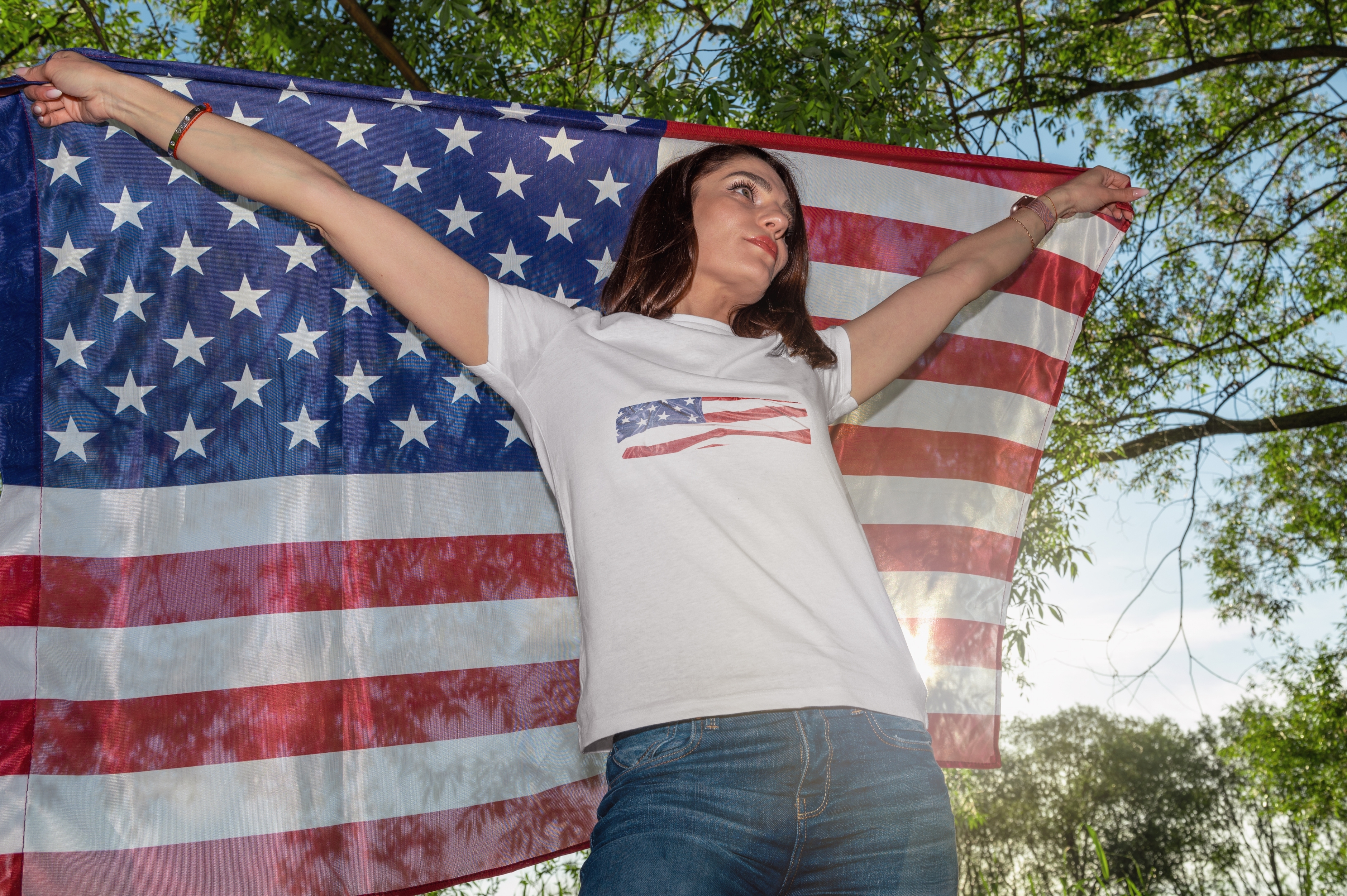 Person holding U.S. flag wearing a T-shirt with a flag design, outdoors under trees, suggesting a patriotic theme