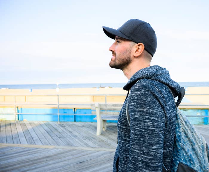 A person wearing a cap and hoodie stands on a boardwalk by the ocean, looking thoughtfully at the sea