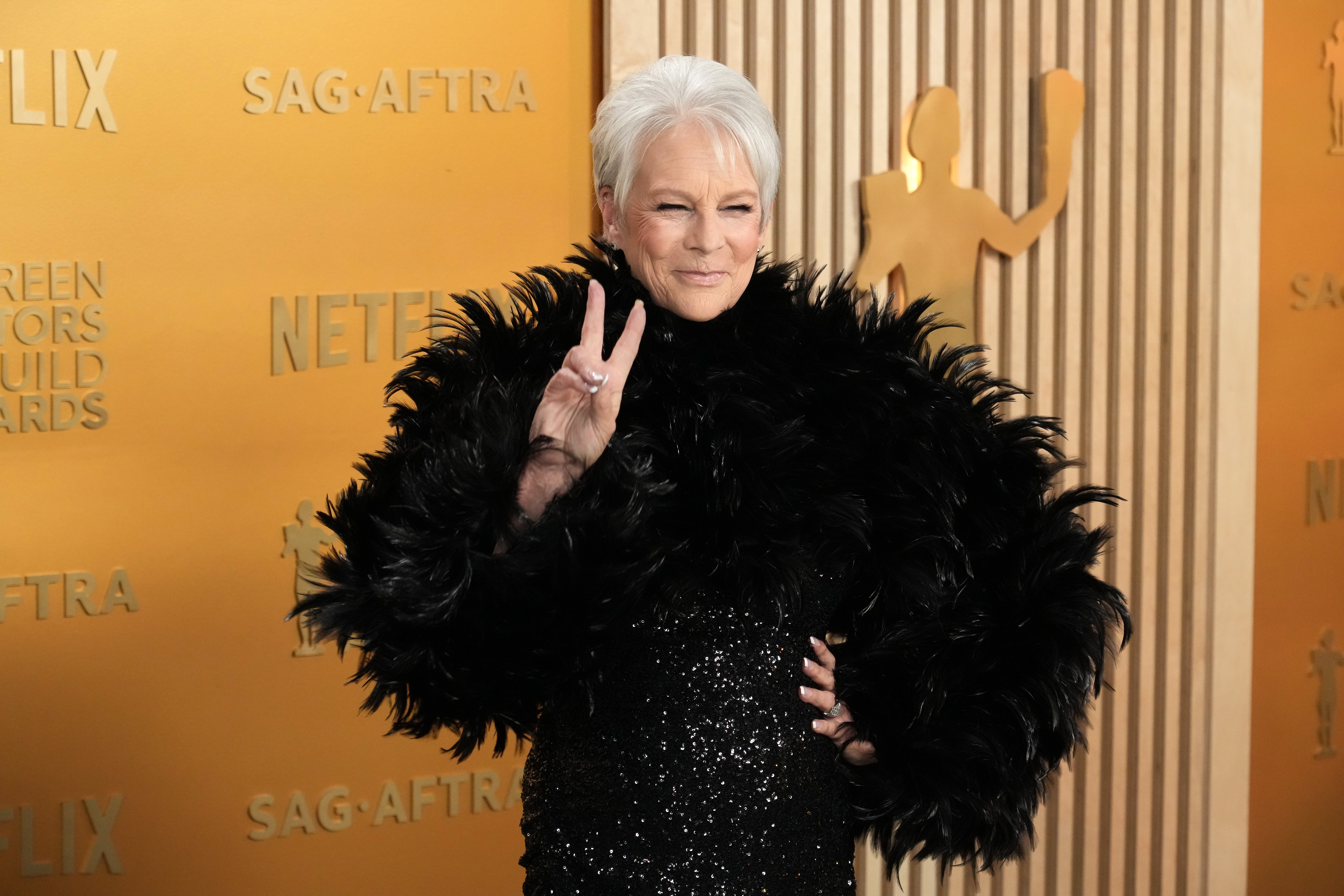 A person in a sequined gown with a dramatic feathered shawl poses on a red carpet, holding up a peace sign