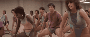 A group of people, dressed in '80s aerobics attire, exercise in a gym class with an energetic atmosphere