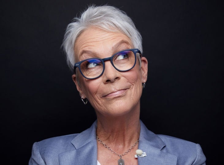 Person with short hair and glasses, wearing a blazer and white shirt, posing with arms crossed against a dark background