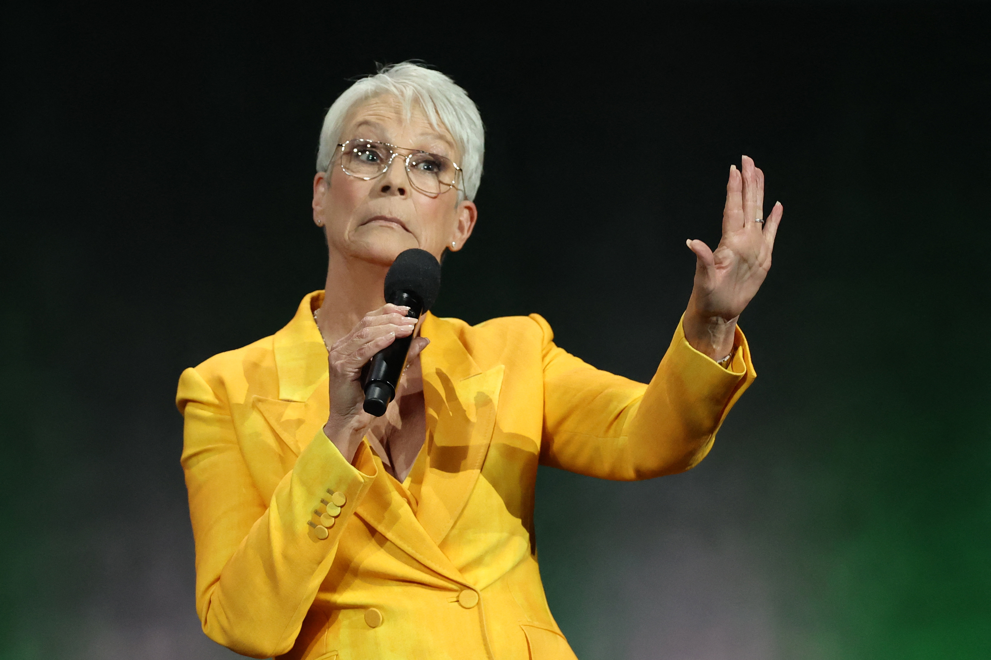 Person on stage, holding a microphone, wearing a tailored yellow suit, gestures with one hand