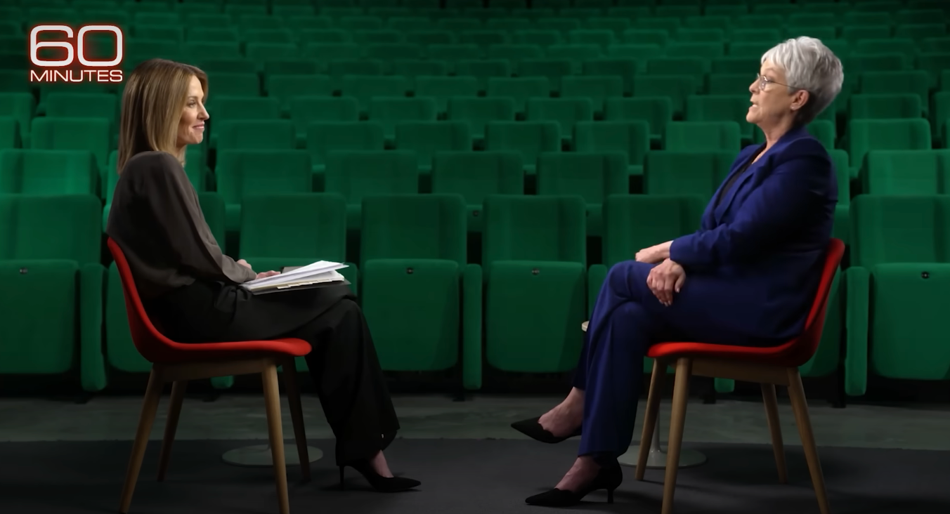 Two people are seated facing each other in a theater setting during a "60 Minutes" interview. One person wears a dark outfit, the other a blue suit