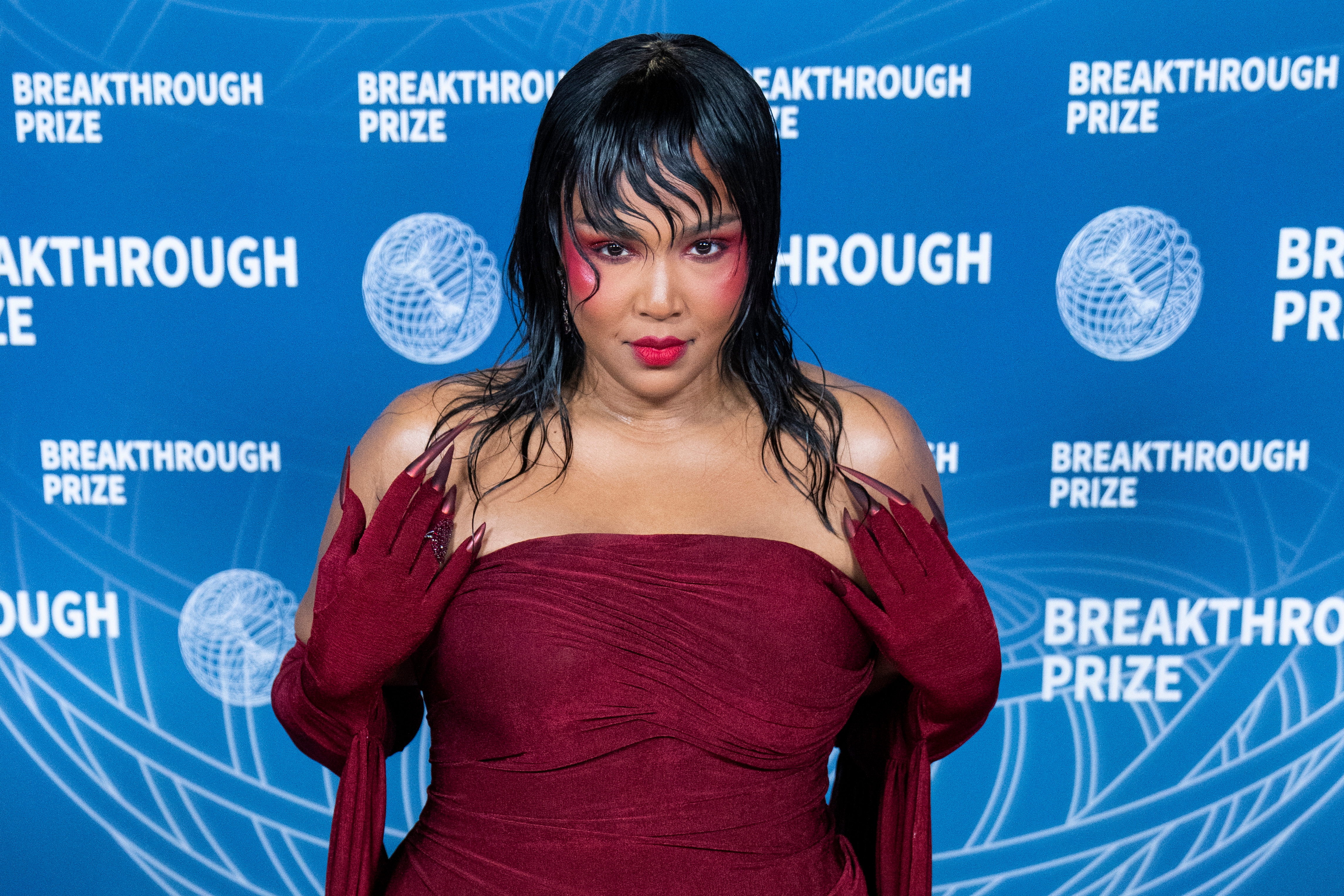 Lizzo on a red carpet in elegant strapless gown with dramatic eye makeup and long gloves, standing against a backdrop with &quot;Breakthrough Prize&quot; text