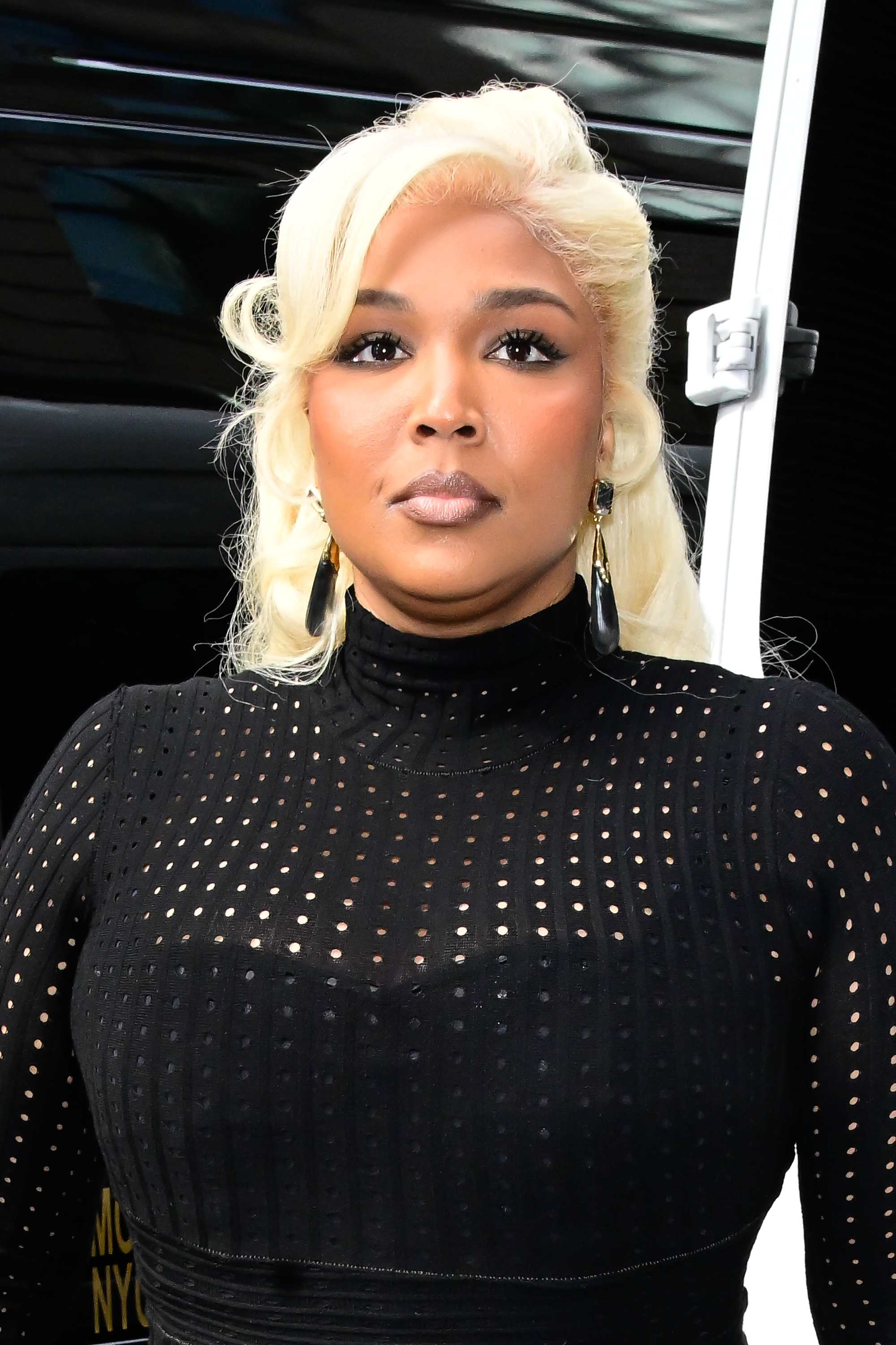 Lizzo with long hair and hoop earrings wearing a black, high-neck, perforated top