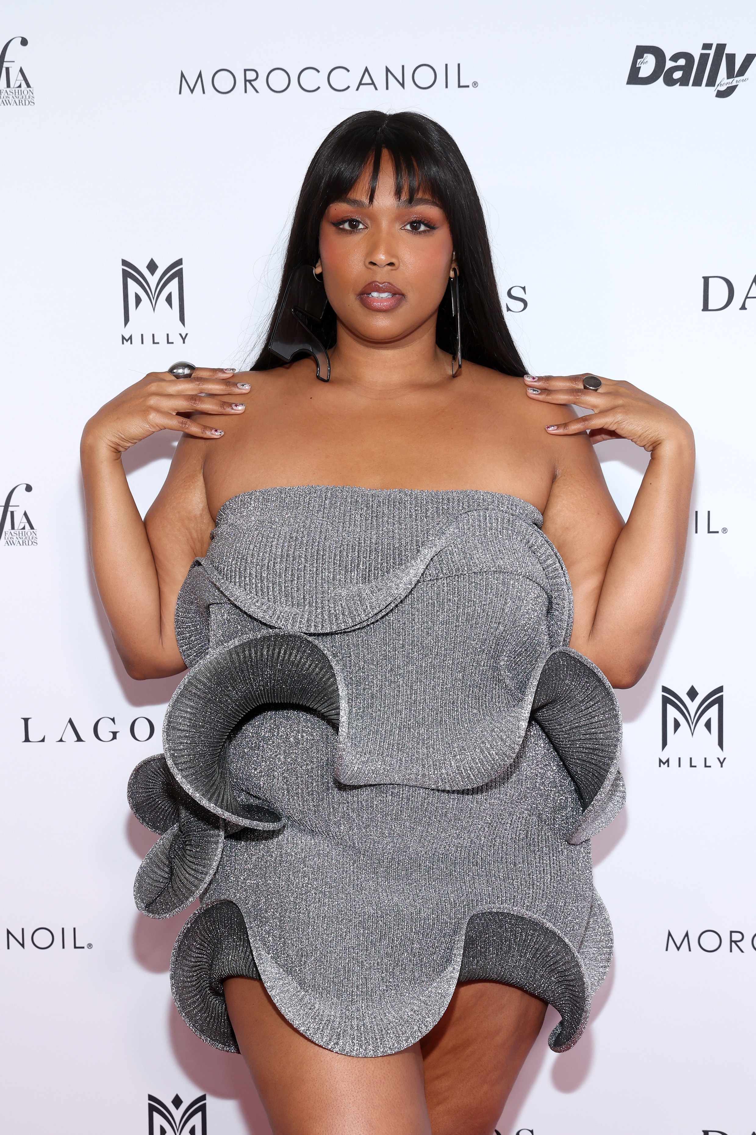 Lizzo on red carpet wearing a unique sculptural dress with wavy elements, posing with hands near face