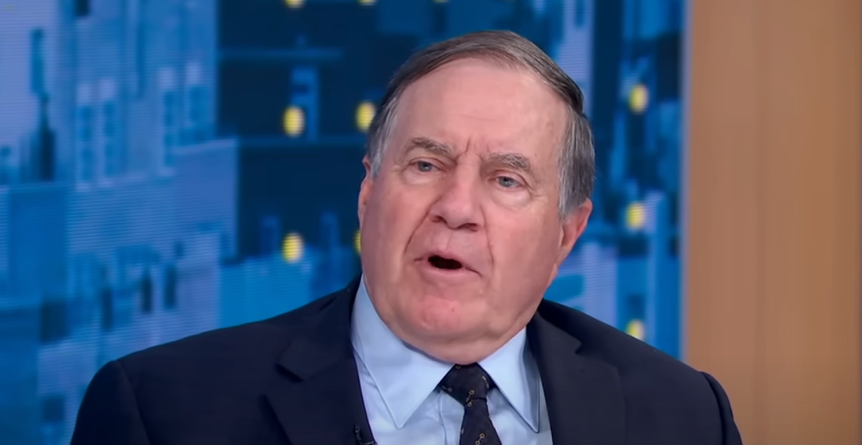 Person speaking on a TV news program, wearing a suit and tie, with a cityscape backdrop