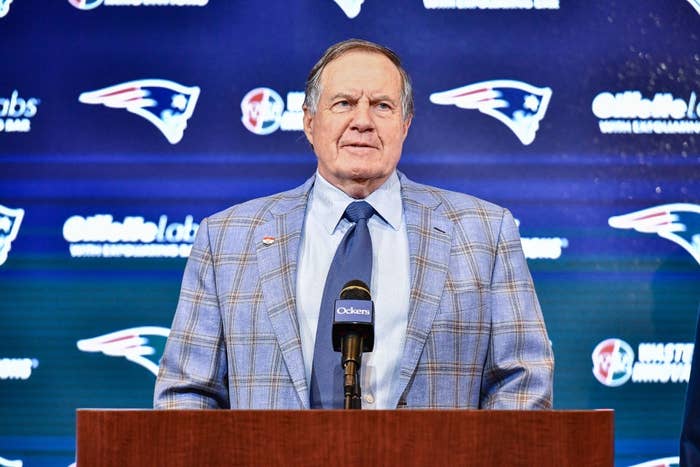 Person in a plaid suit and tie stands at a podium with sports logos in the background