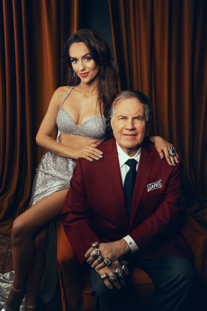Woman in sparkling gown poses behind a seated man in red blazer with multiple champion rings, in front of draped curtains