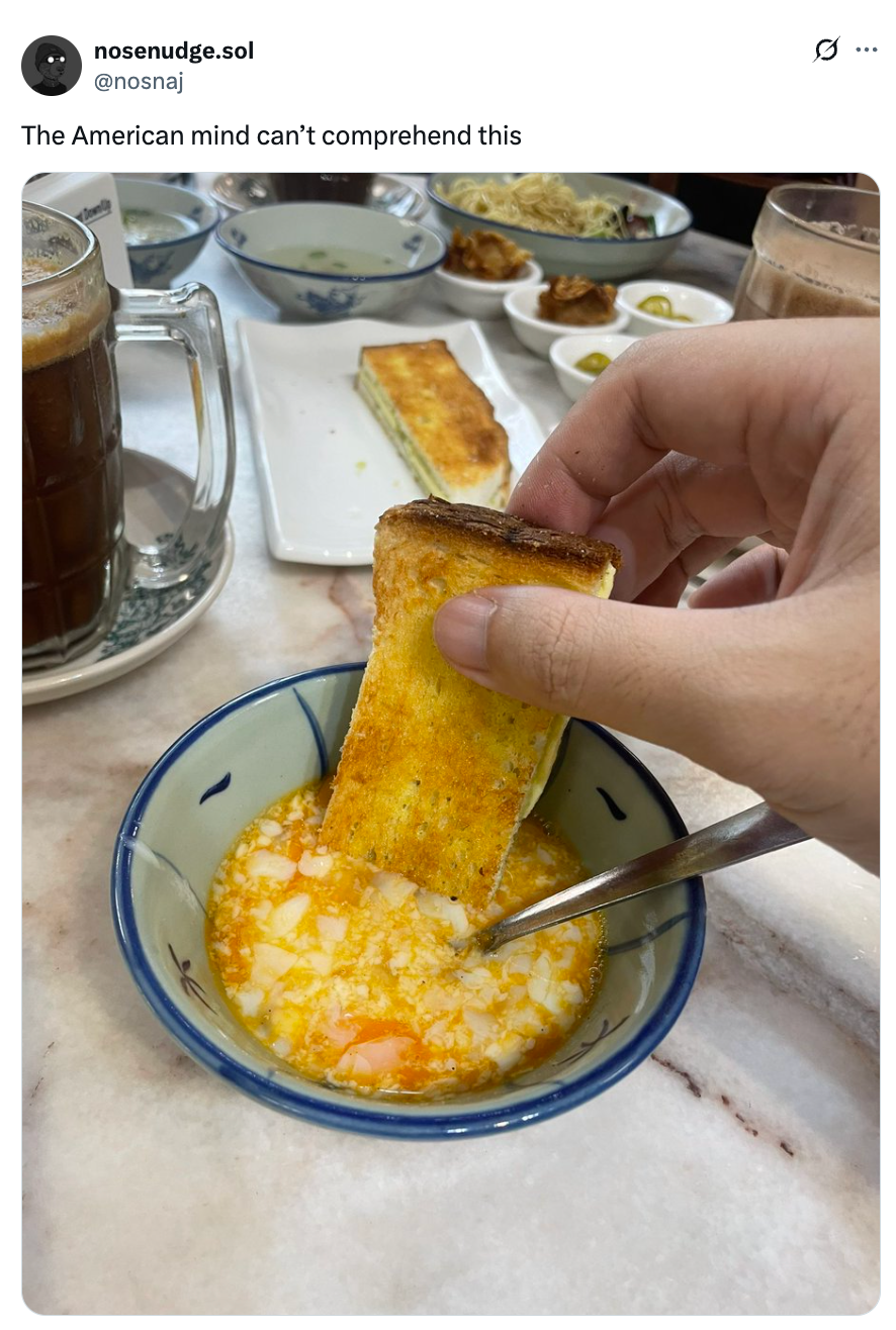 A hand dips toast into a bowl of soup or porridge, with various dishes and a drink in the background. Text reads: "The American mind can’t comprehend this."