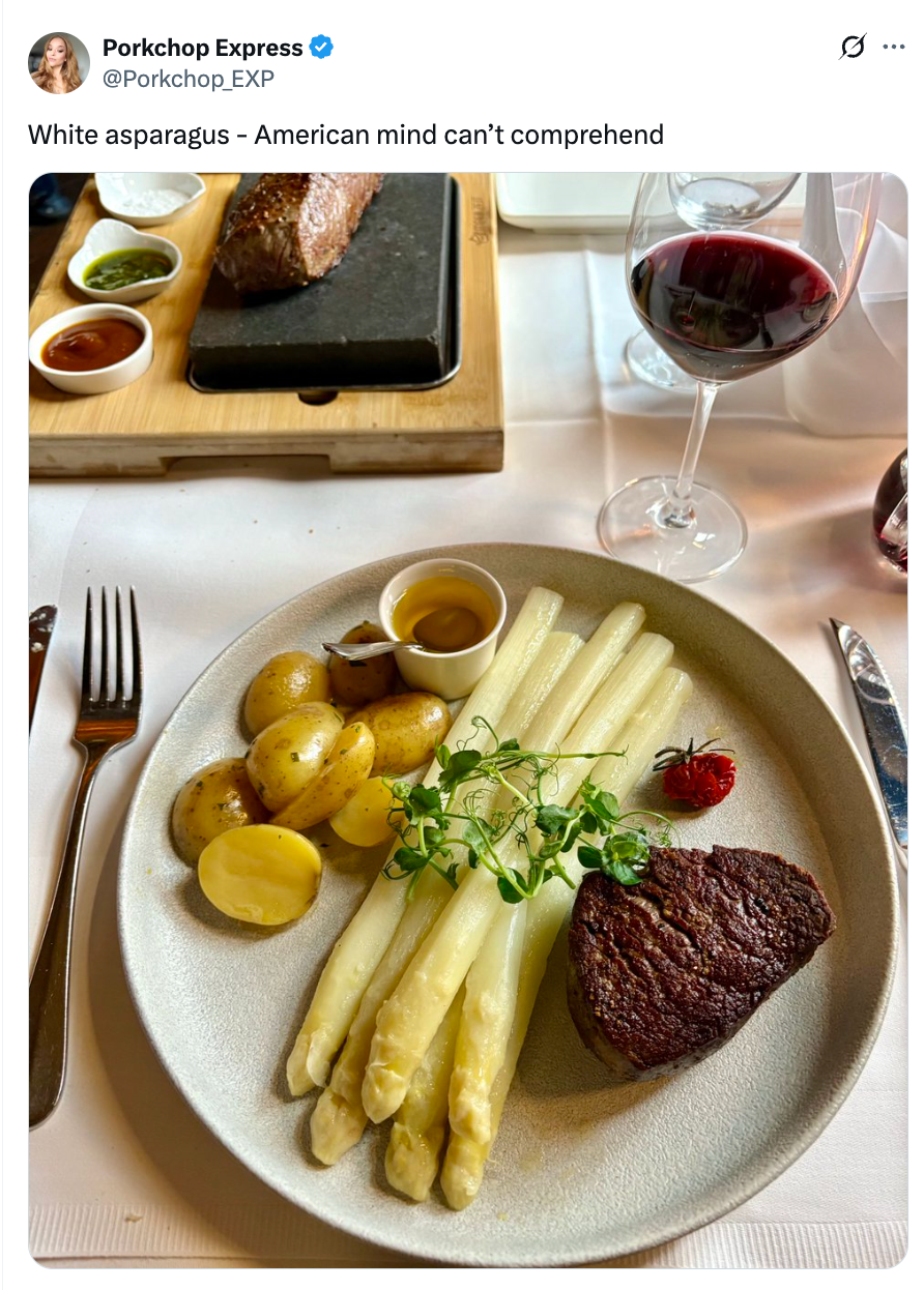Plate with white asparagus, steak, and potatoes. A glass of red wine and sauces are on the table. Tweet text: "White asparagus – American mind can't comprehend."