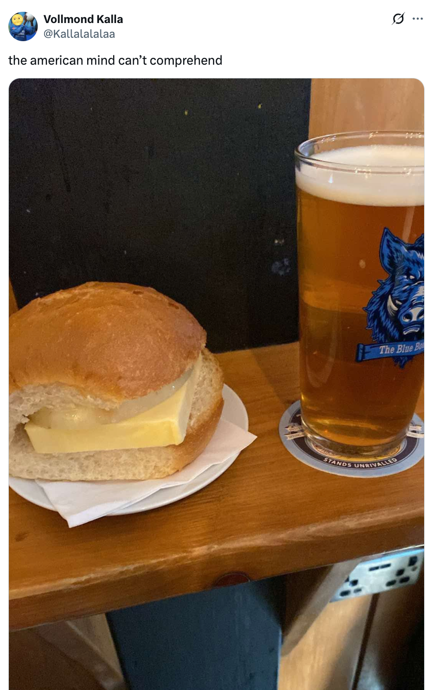 A slice of cheese sandwiched in a bun next to a pint of beer on a wooden table