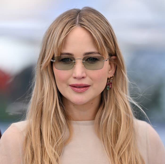 Jennifer Lawrence in a sophisticated outfit and sunglasses, standing outdoors at a formal event. They have long hair and are wearing earrings