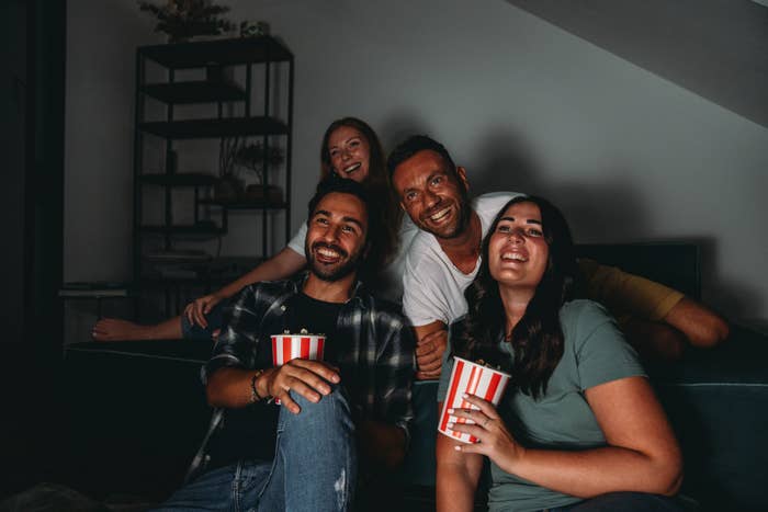 Four friends smiling and sitting on a couch, holding striped popcorn containers, watching a movie