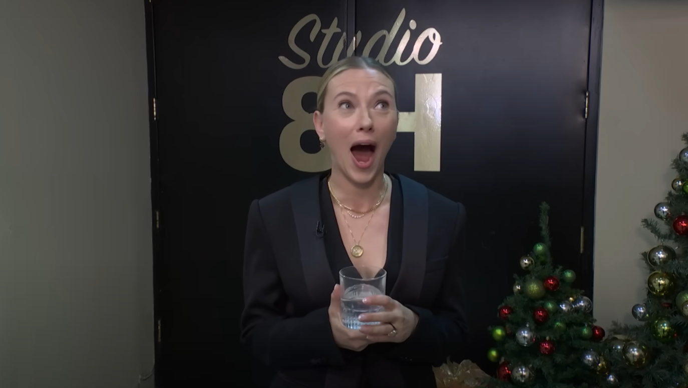 Person in black blazer holding a glass of water, appearing surprised or excited, stands in front of Studio 8H sign with Christmas decorations nearby