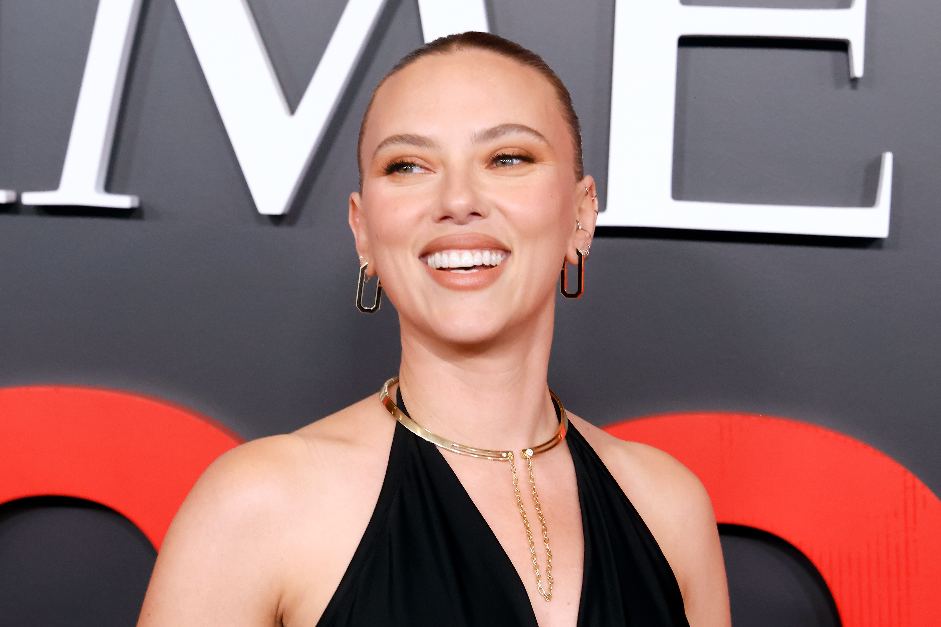 Person smiling on a red carpet, wearing a sleeveless black gown and gold jewelry