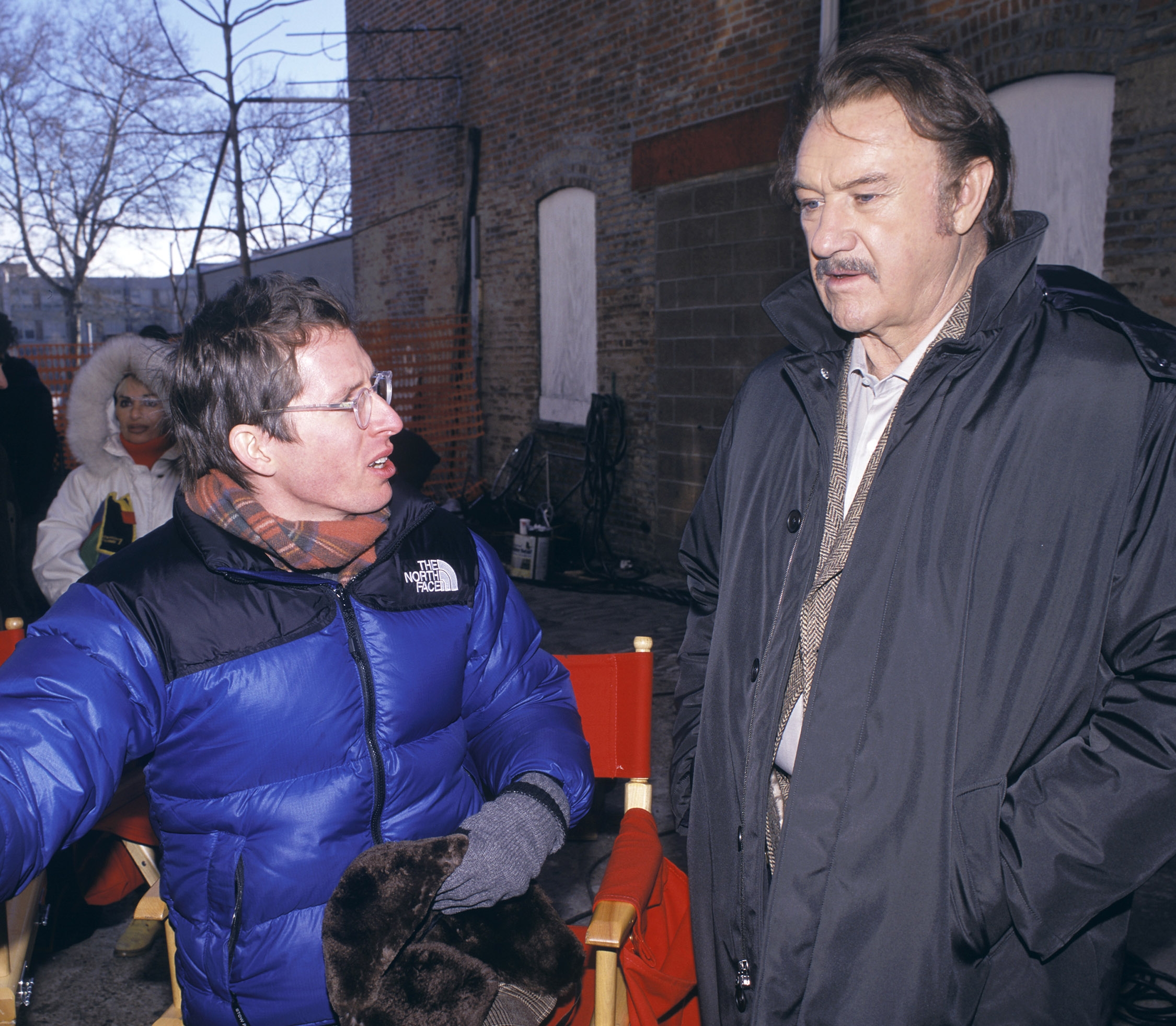 Two men outside talking; one in a blue winter jacket and glasses, the other in a long dark coat with a mustache
