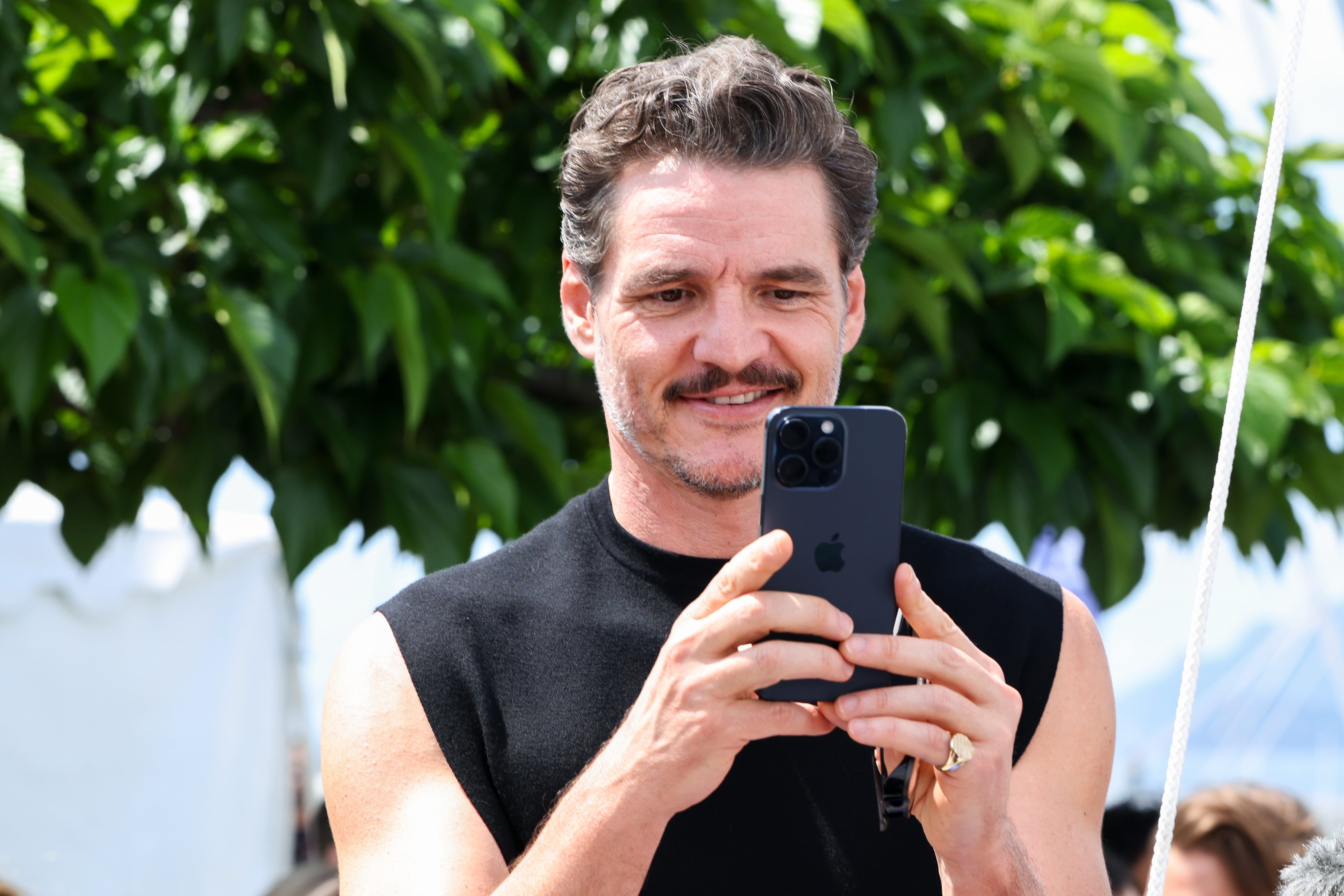 Person outdoors, looking at phone with a smile. They're wearing a sleeveless top and standing near lush tree foliage