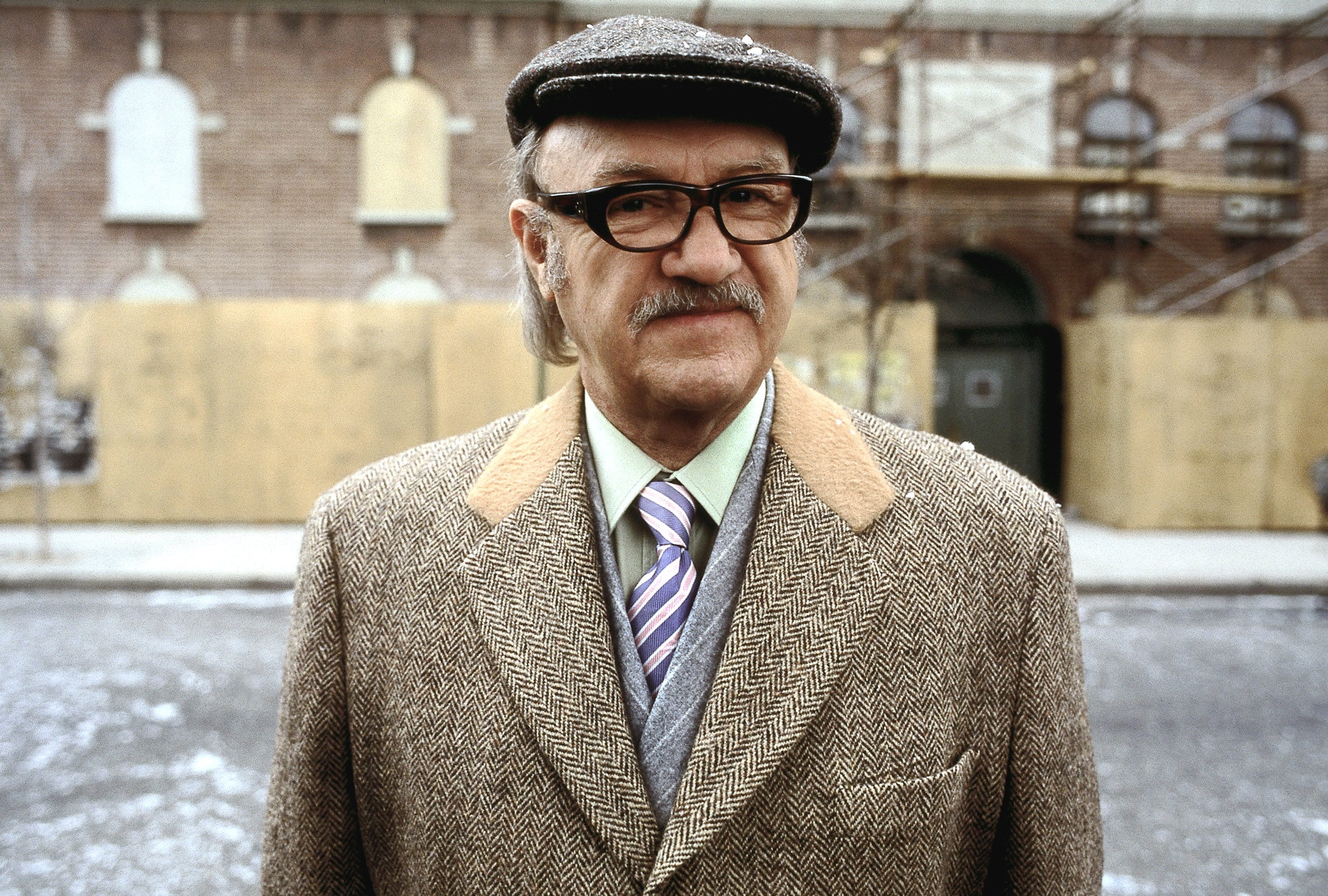 Gene Hackman wearing a herringbone blazer, flat cap, and striped tie stands outdoors on a snowy street with a brick building in the background