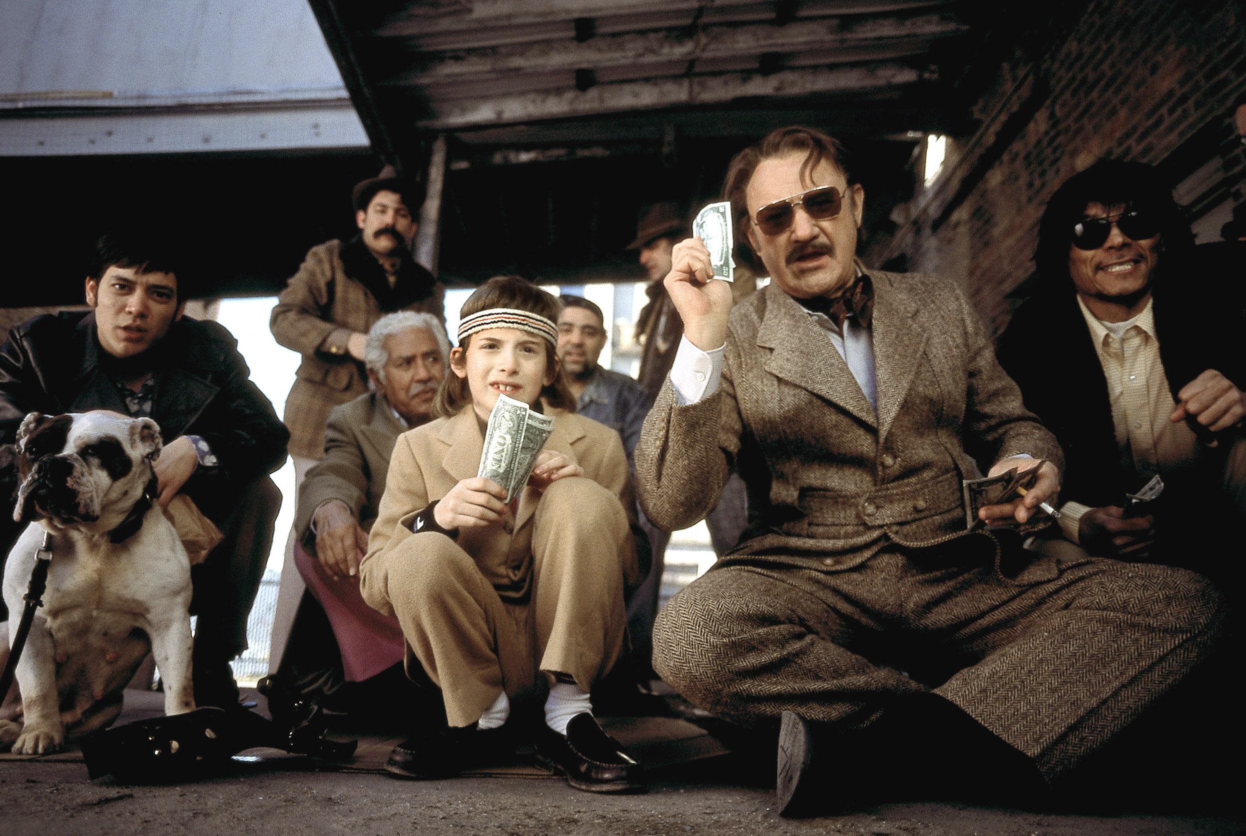 Group of men, including a child, sitting with $1 bills; a bulldog is beside them. They are dressed in vintage-style clothing and appear to gamble outdoors